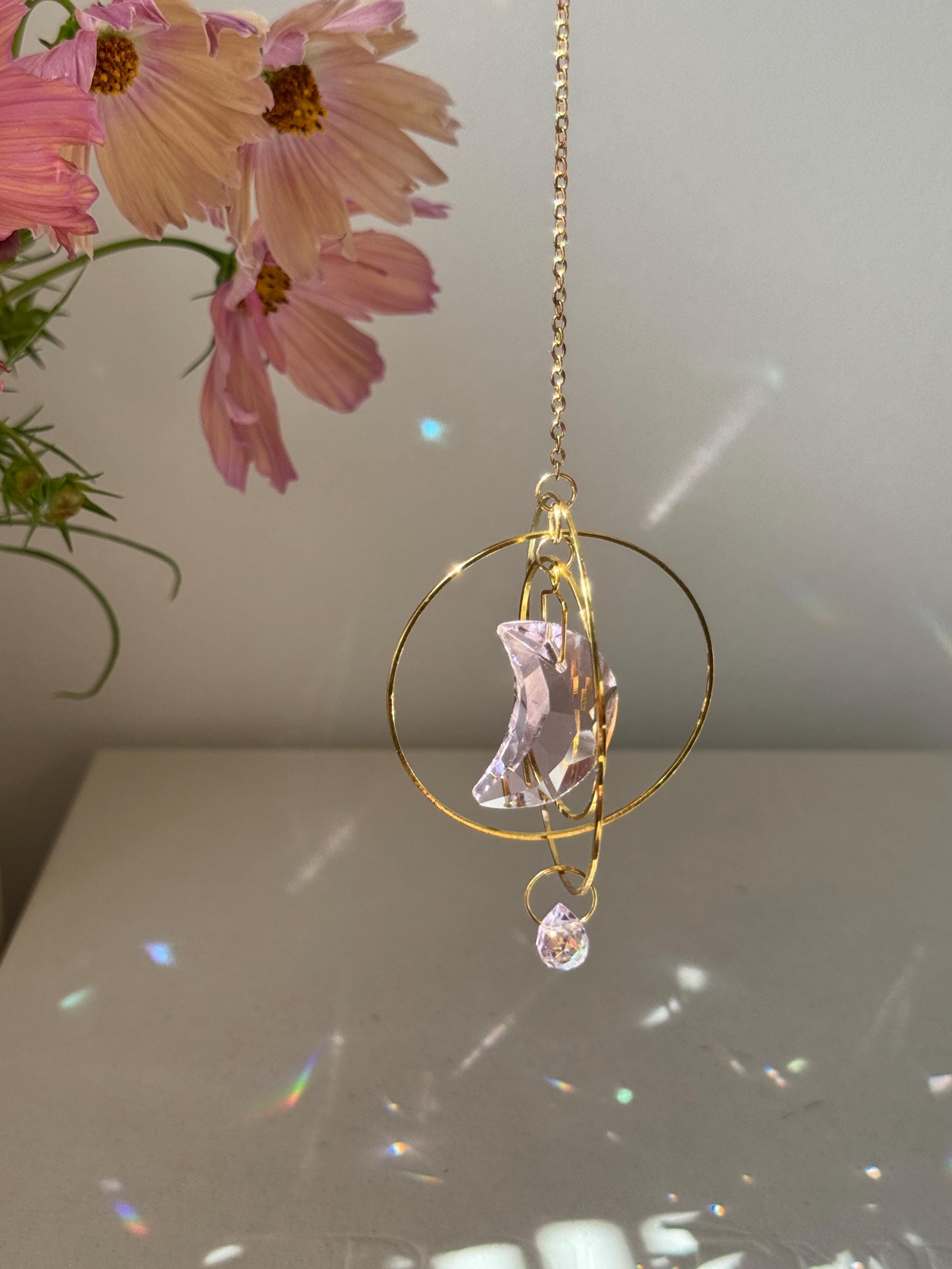 Handmade pink crescent moon suncatcher hanging in a sunny window creating rainbow prisms on the wall