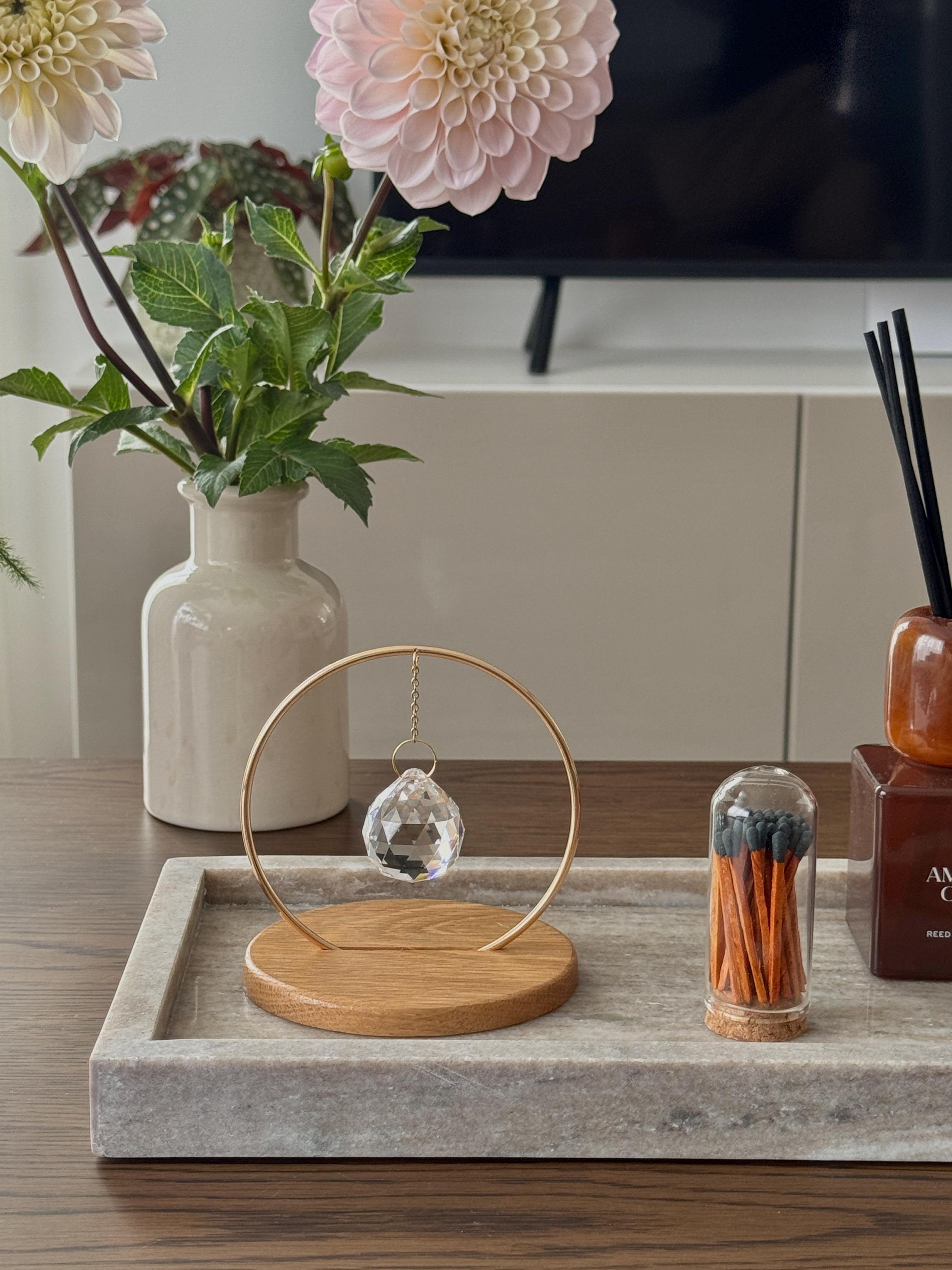 A clear, faceted crystal prism sphere hangs by a delicate gold chain within a circular brass frame, titled "The Circle." The suncatcher is mounted on a smooth, light oak wooden base. Sunlight passes through the crystal, scattering vibrant rainbow light leaks and spectrum dots across a white background and onto gnarled, aesthetic tree branches. The scene is bright, minimalist, and serene.