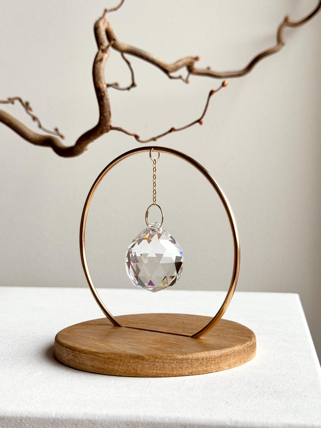 A clear, faceted crystal prism sphere hangs by a delicate gold chain within a circular brass frame, titled "The Circle." The suncatcher is mounted on a smooth, light oak wooden base. Sunlight passes through the crystal, scattering vibrant rainbow light leaks and spectrum dots across a white background and onto gnarled, aesthetic tree branches. The scene is bright, minimalist, and serene.