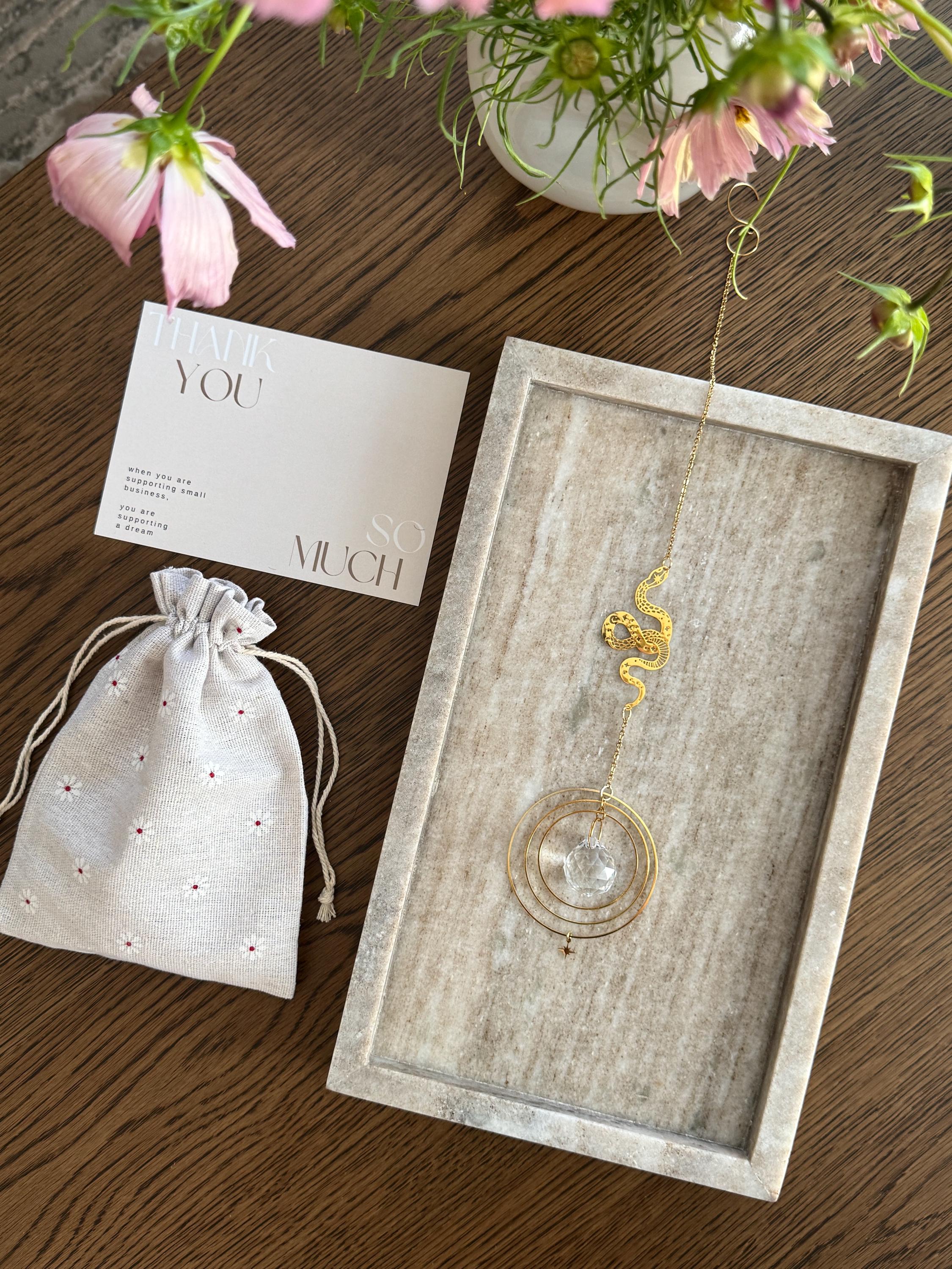 Decorative glass pendant with drawstring linen bag and card on a wooden surface with flowers.