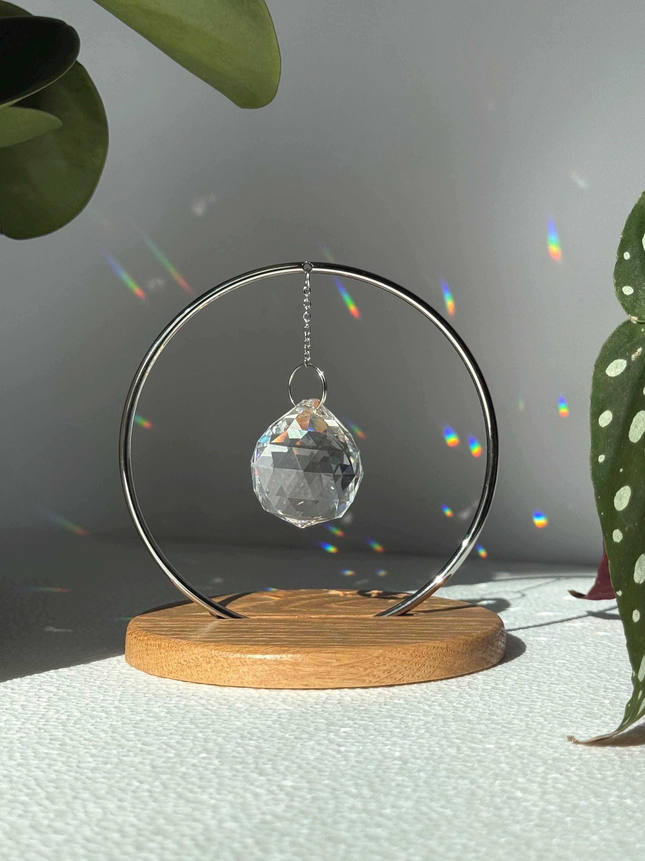 Minimalist "The Circle" tabletop suncatcher featuring a faceted crystal prism hanging inside a stainless steel ring on a round American oak base, casting vibrant rainbows on a white wall.