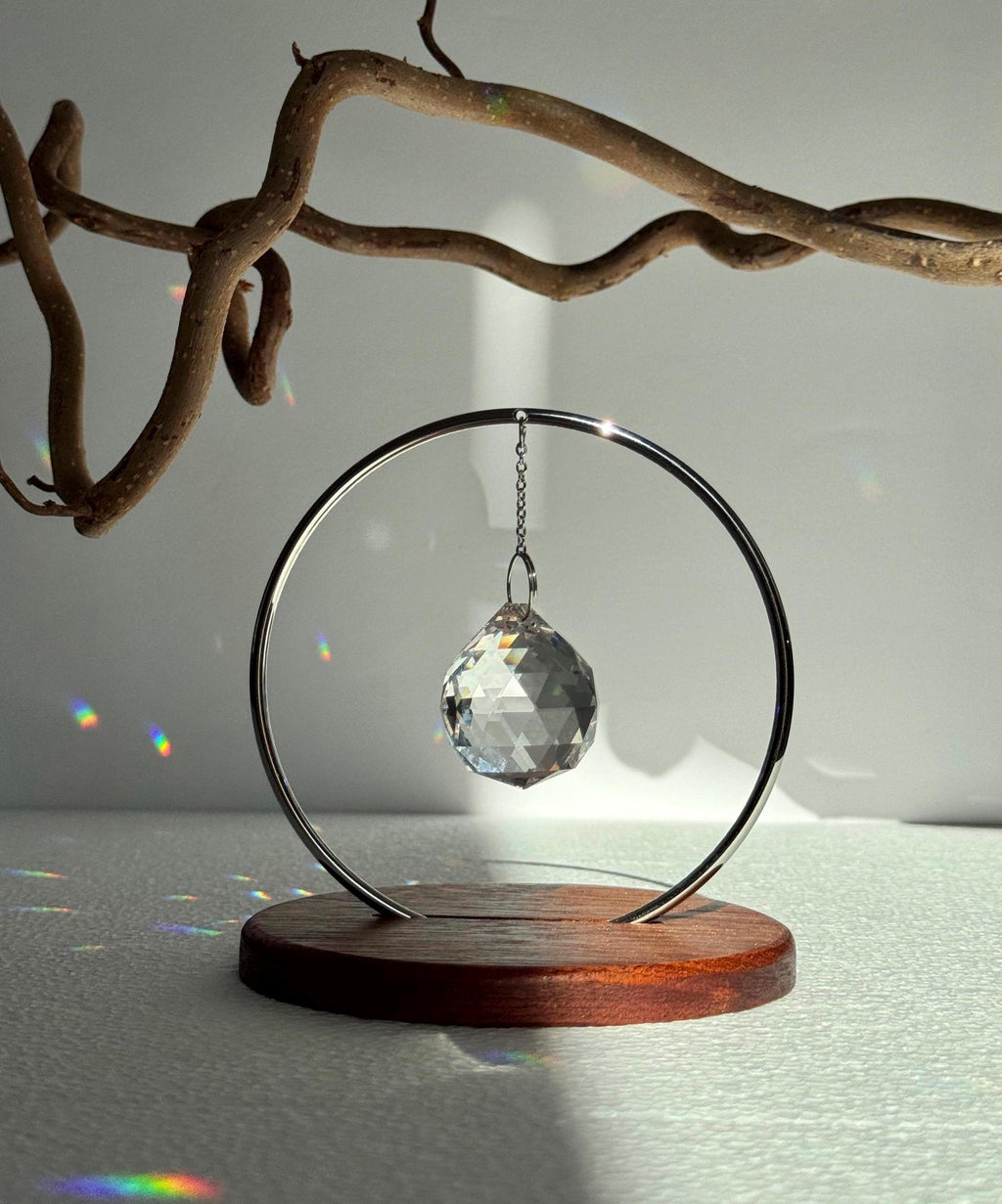 The Circle tabletop suncatcher featuring a silver stainless steel ring and faceted crystal prism casting vibrant rainbows on a white surface.