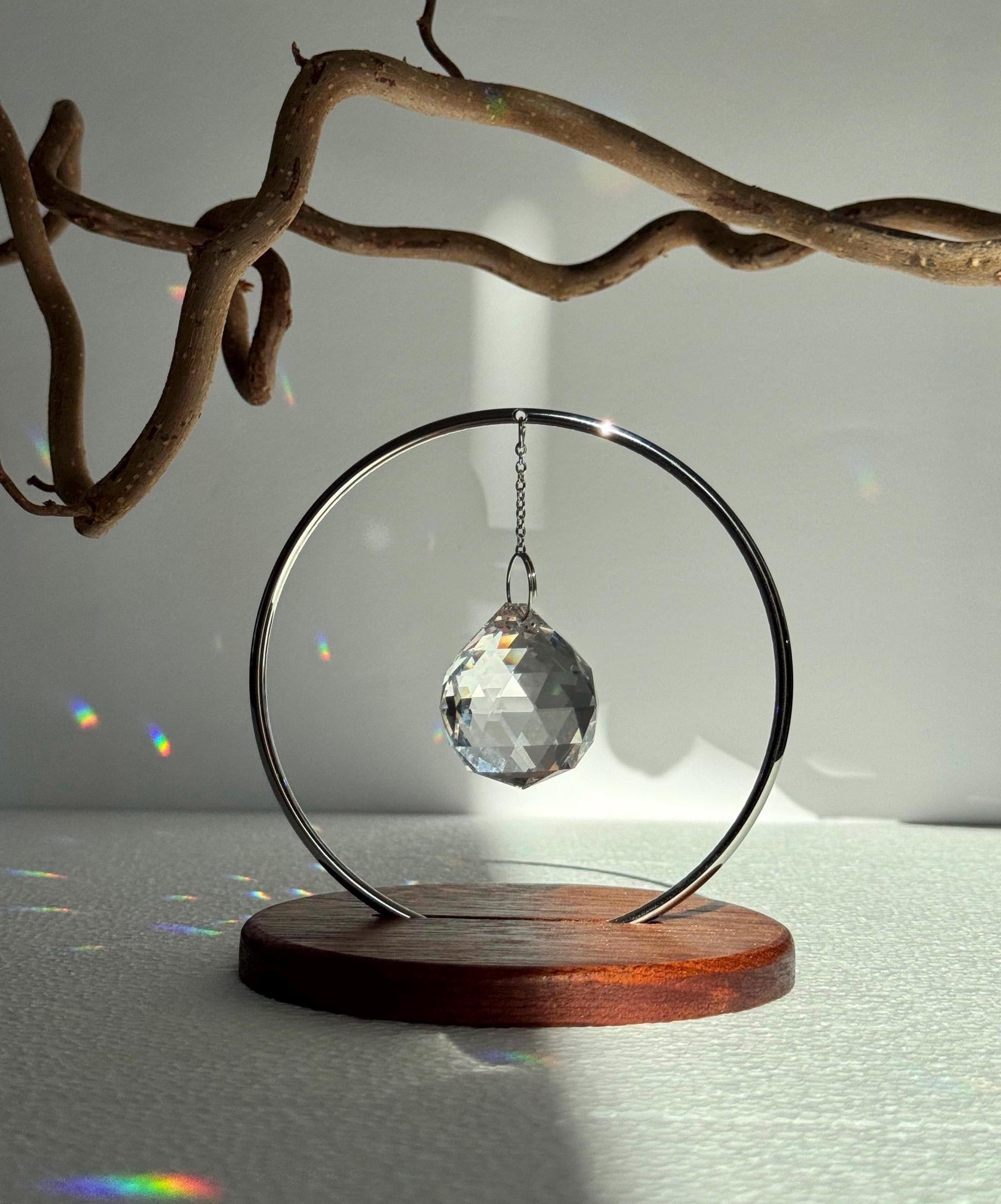 The Circle tabletop suncatcher featuring a silver stainless steel ring and faceted crystal prism casting vibrant rainbows on a white surface.