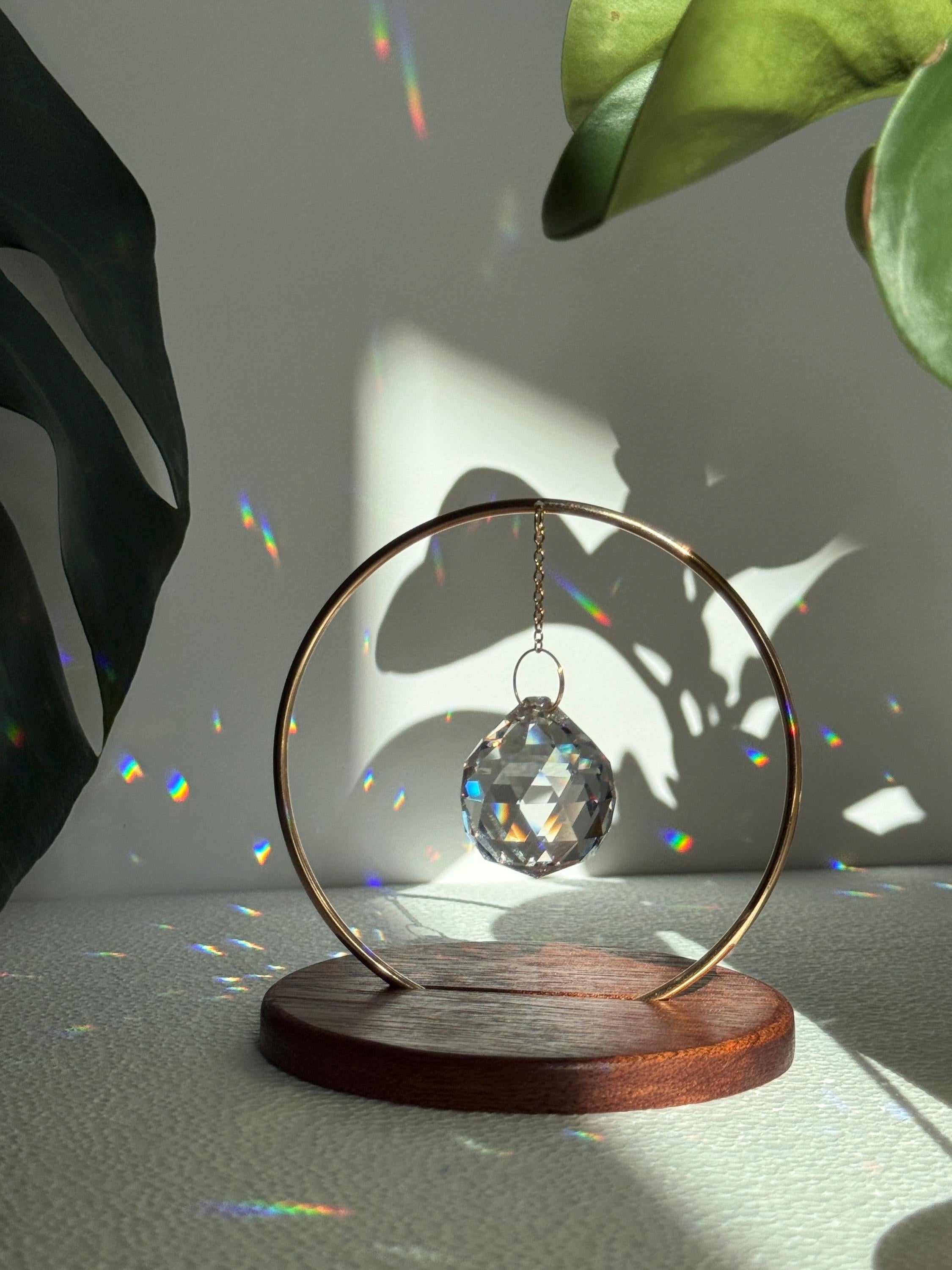 The Circle - A minimalist Sonnenfänger (suncatcher) featuring a high-quality faceted Kristall-Prisma suspended by a delicate gold stainless steel chain within a matching gold ring. The modern tabletop design rests on a solid, dark redwood base (Echtholz-Sockel) and is shown creating vibrant rainbow light reflections (Regenbogen-Effekt) across the background. This elegant Fensterdeko is perfect as a mindful Geschenkidee for home or office.