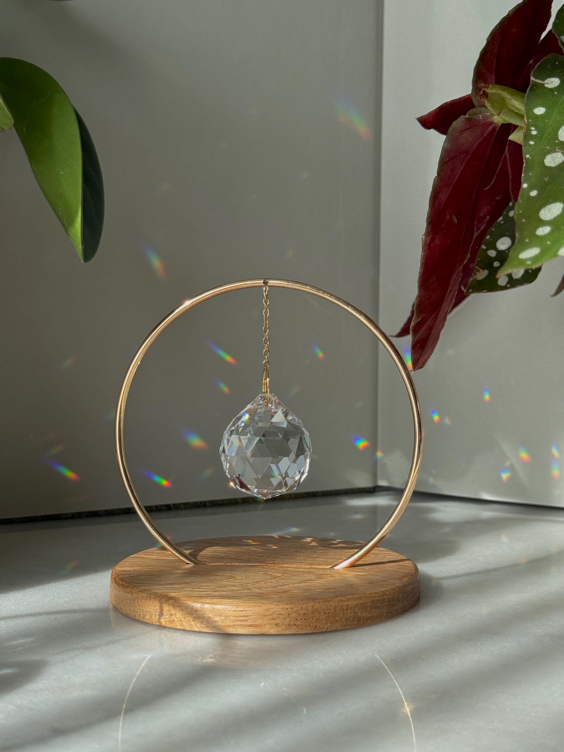 A clear, faceted crystal prism sphere hangs by a delicate gold chain within a circular brass frame, titled "The Circle." The suncatcher is mounted on a smooth, light oak wooden base. Sunlight passes through the crystal, scattering vibrant rainbow light leaks and spectrum dots across a white background and onto gnarled, aesthetic tree branches. The scene is bright, minimalist, and serene.