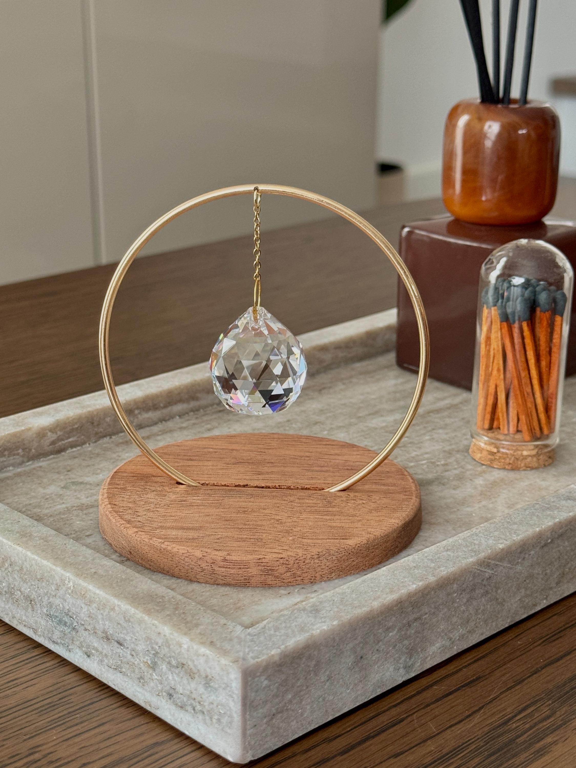 The Circle - A minimalist Sonnenfänger (suncatcher) featuring a high-quality faceted Kristall-Prisma suspended by a delicate gold stainless steel chain within a matching gold ring. The modern tabletop design rests on a solid, dark redwood base (Echtholz-Sockel) and is shown creating vibrant rainbow light reflections (Regenbogen-Effekt) across the background. This elegant Fensterdeko is perfect as a mindful Geschenkidee for home or office.