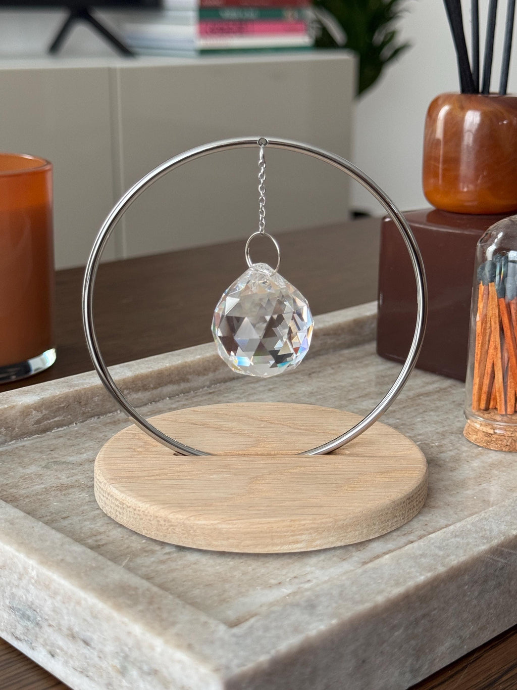 Minimalist "The Circle" tabletop suncatcher featuring a faceted crystal prism hanging inside a stainless steel ring on a round American oak base, casting vibrant rainbows on a white wall.