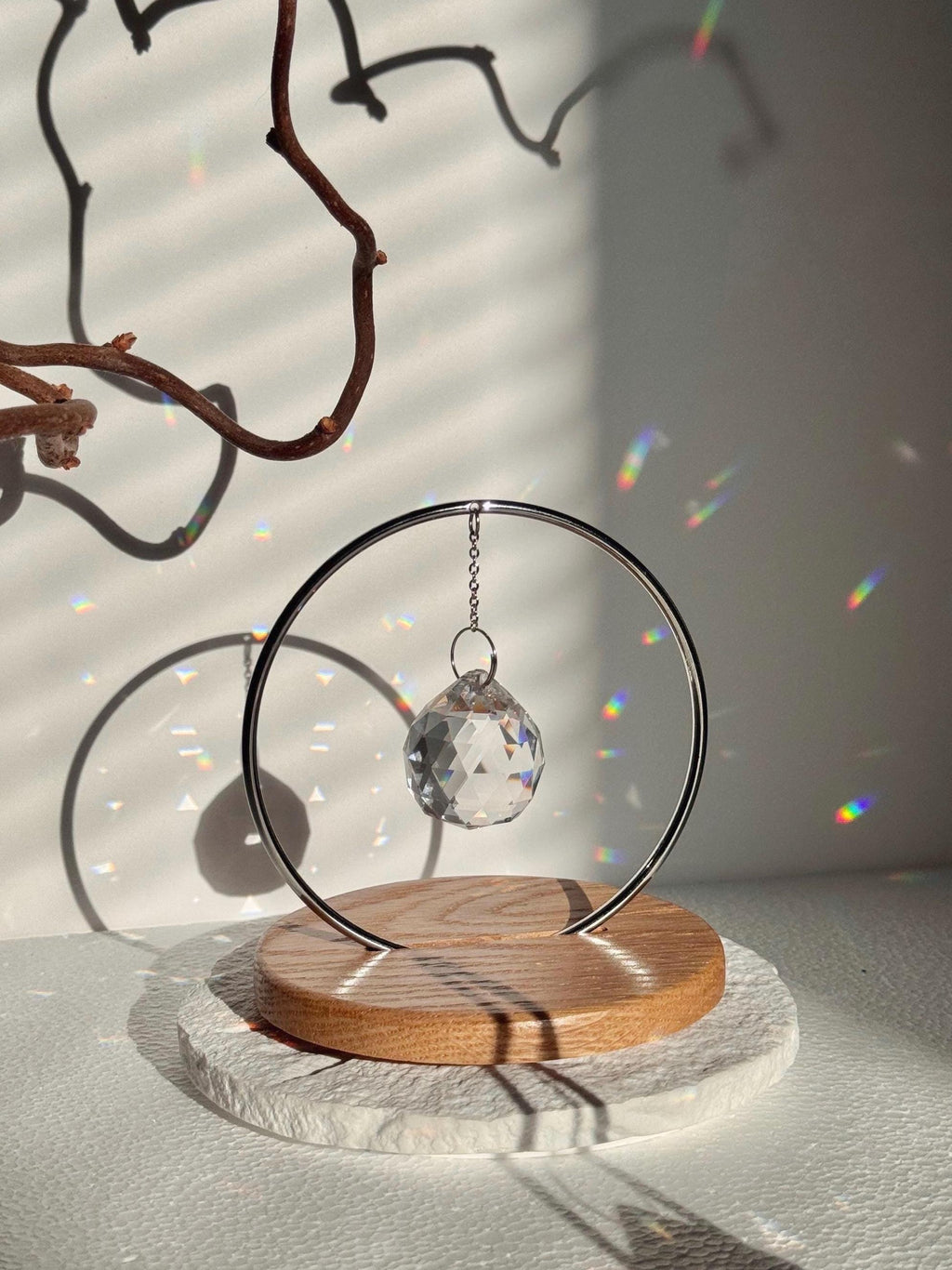Minimalist "The Circle" tabletop suncatcher featuring a faceted crystal prism hanging inside a stainless steel ring on a round American oak base, casting vibrant rainbows on a white wall.