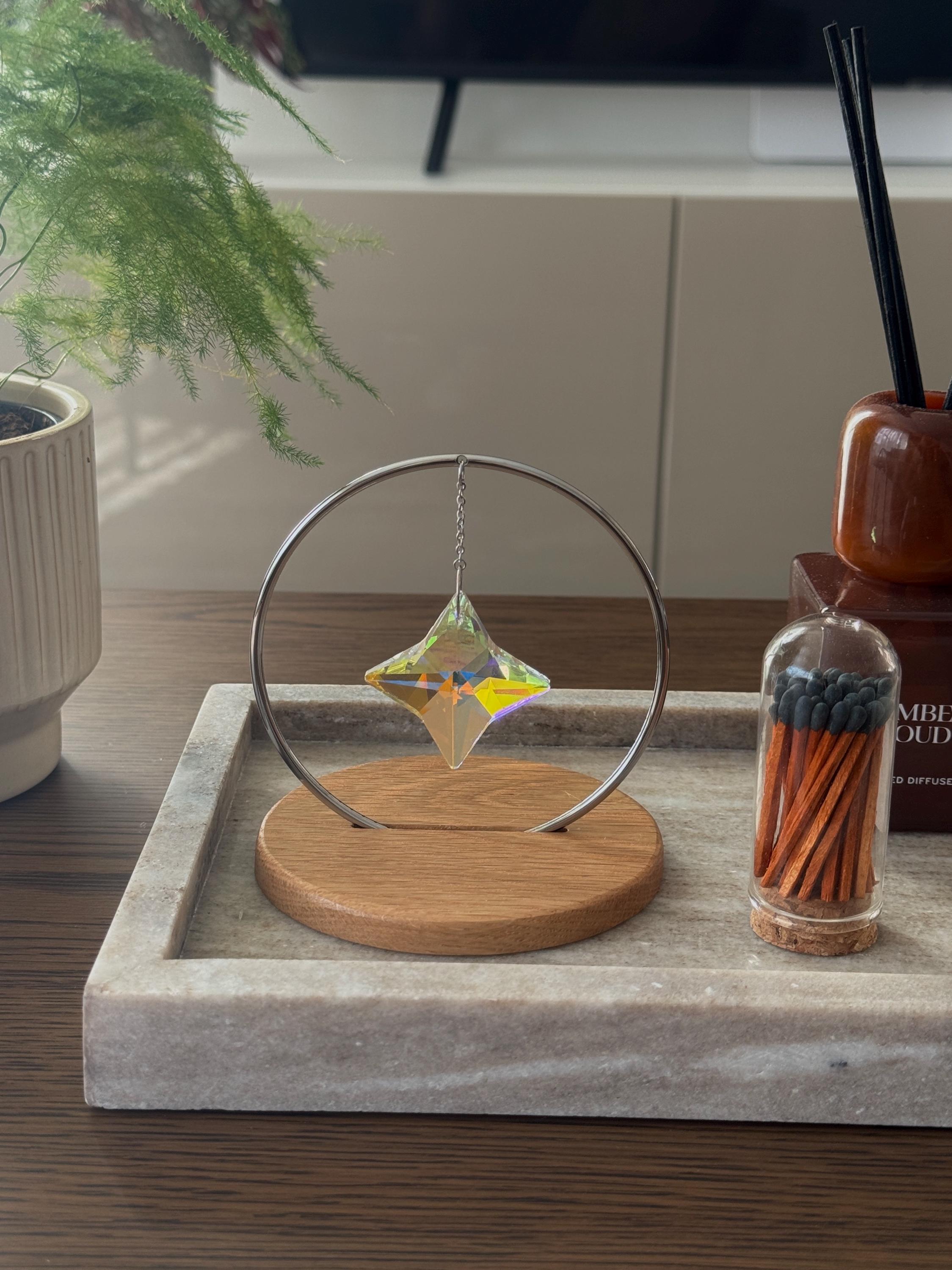 A minimalist Heim decor piece: The Halo Suncatcher featuring a brilliant Kristall-Stern (crystal star) suspended in a silver ring. The star catches the light, scattering vibrant rainbows across a white surface next to a warm wooden base. Wunderschön!