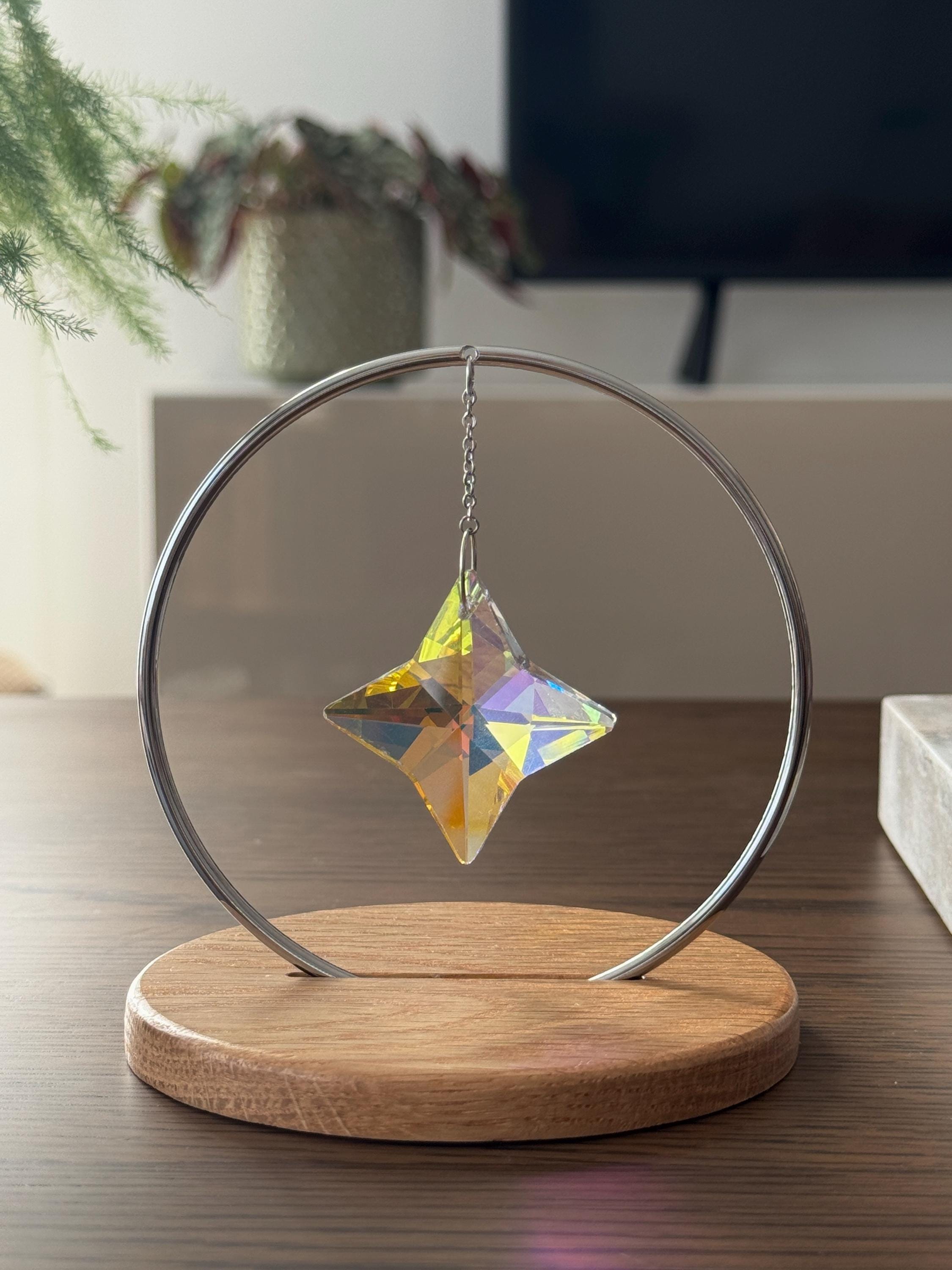 A minimalist Heim decor piece: The Halo Suncatcher featuring a brilliant Kristall-Stern (crystal star) suspended in a silver ring. The star catches the light, scattering vibrant rainbows across a white surface next to a warm wooden base. Wunderschön!