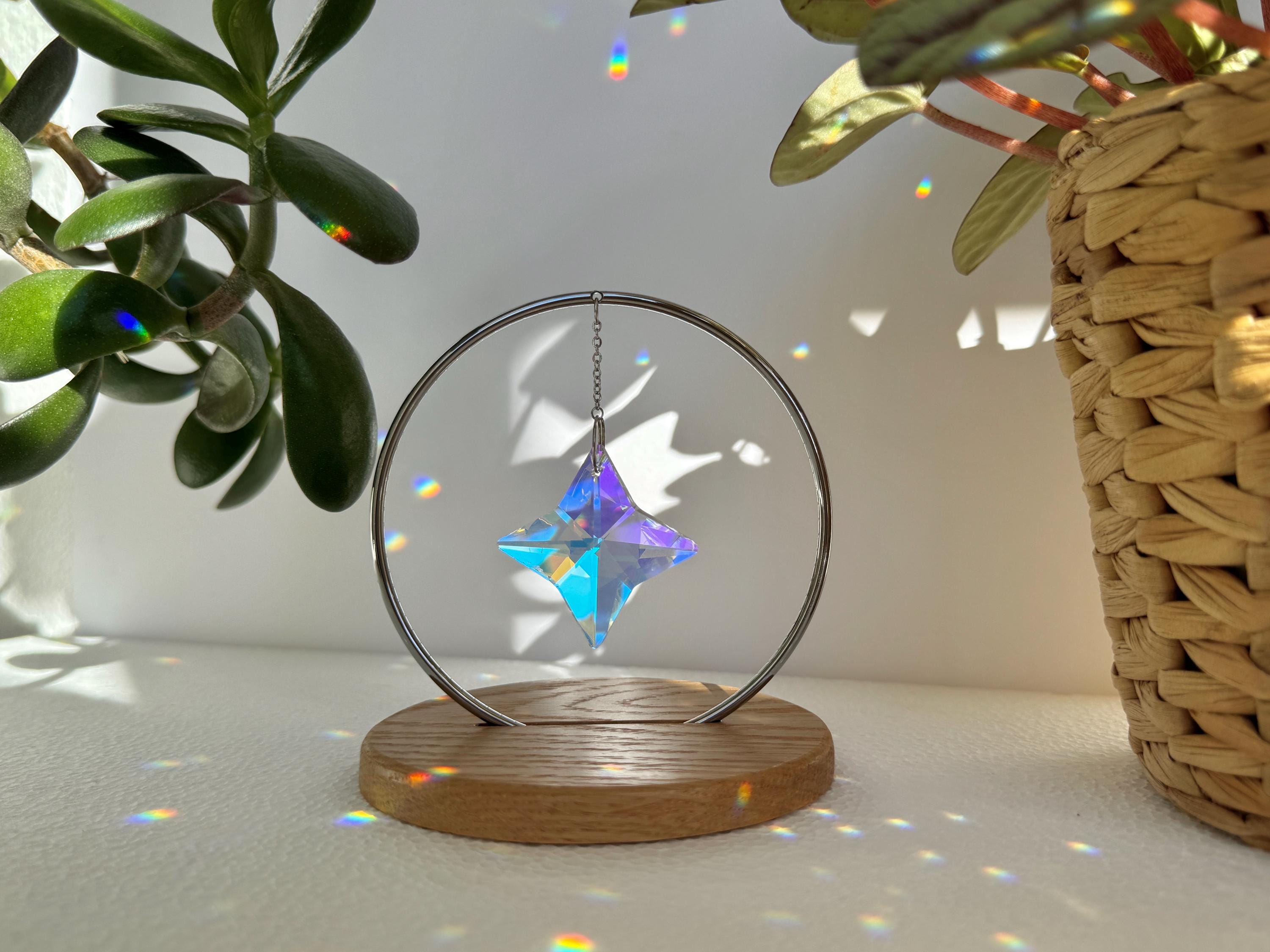 A minimalist Heim decor piece: The Halo Suncatcher featuring a brilliant Kristall-Stern (crystal star) suspended in a silver ring. The star catches the light, scattering vibrant rainbows across a white surface next to a warm wooden base. Wunderschön!