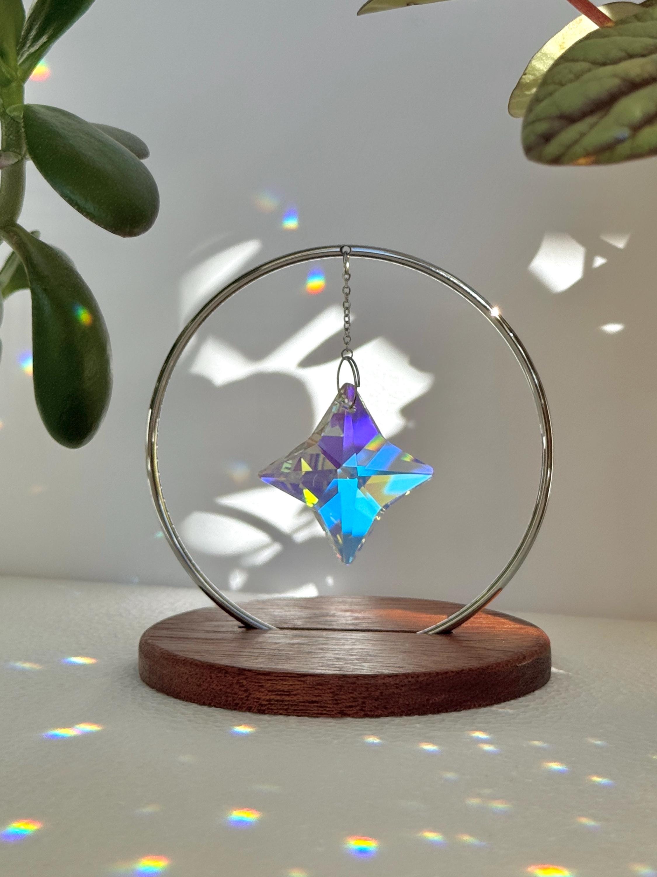 A minimalist Heim decor piece: The Halo Suncatcher featuring a brilliant Kristall-Stern (crystal star) suspended in a silver ring. The star catches the light, scattering vibrant rainbows across a white surface next to a warm wooden base. Wunderschön!
