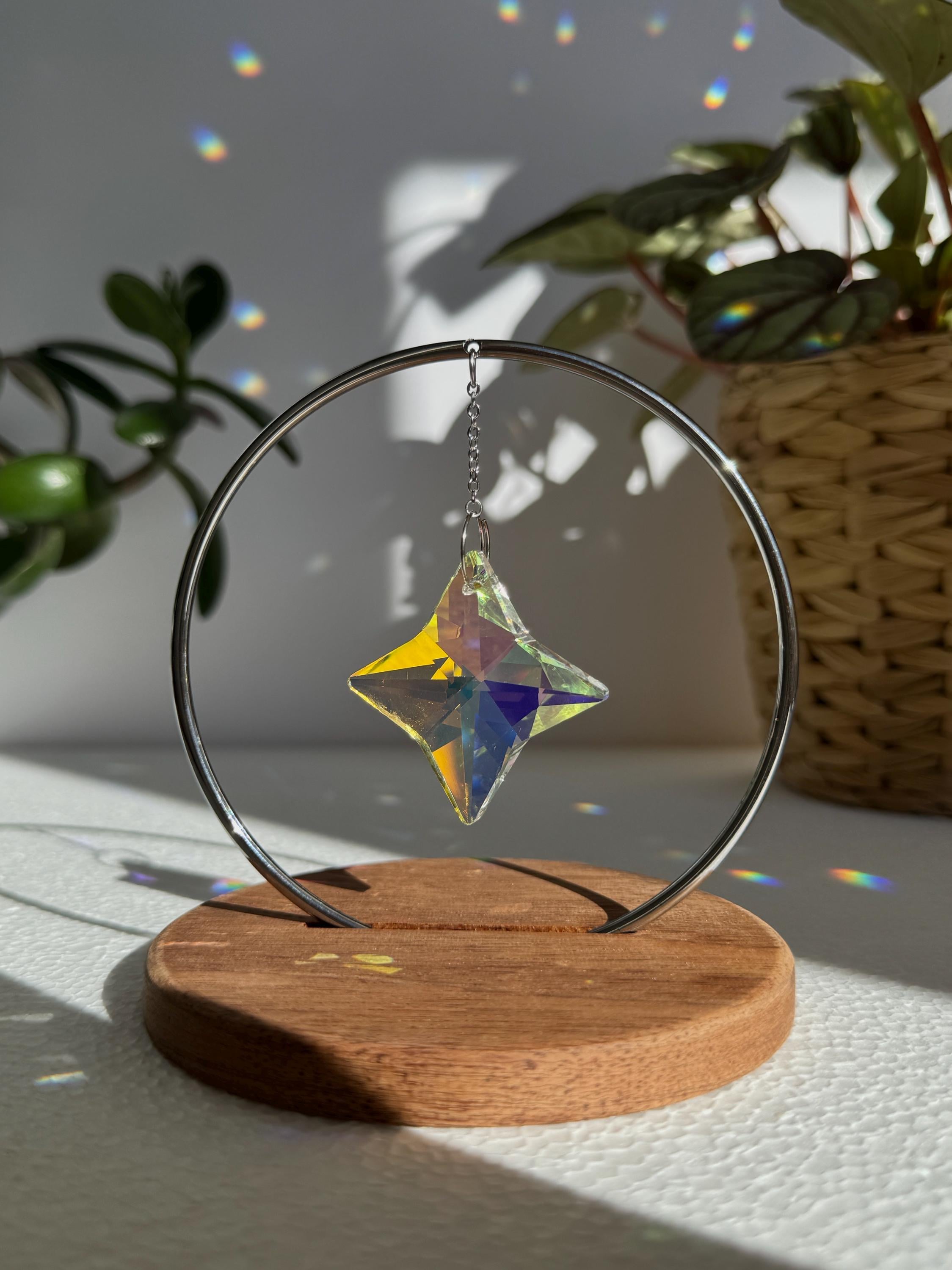 A minimalist Heim decor piece: The Halo Suncatcher featuring a brilliant Kristall-Stern (crystal star) suspended in a silver ring. The star catches the light, scattering vibrant rainbows across a white surface next to a warm wooden base. Wunderschön!