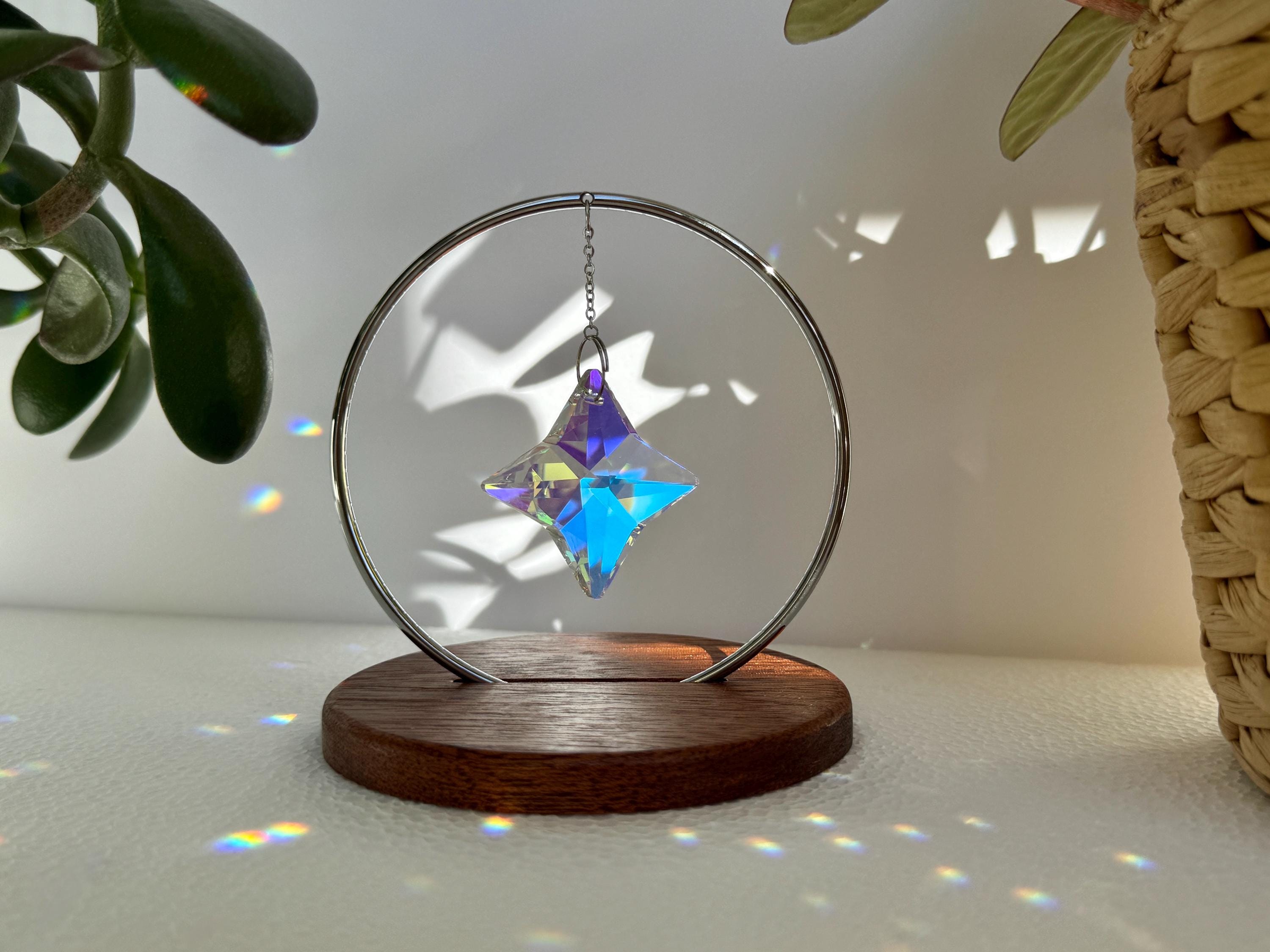 A minimalist Heim decor piece: The Halo Suncatcher featuring a brilliant Kristall-Stern (crystal star) suspended in a silver ring. The star catches the light, scattering vibrant rainbows across a white surface next to a warm wooden base. Wunderschön!