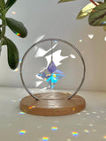 A minimalist Heim decor piece: The Halo Suncatcher featuring a brilliant Kristall-Stern (crystal star) suspended in a silver ring. The star catches the light, scattering vibrant rainbows across a white surface next to a warm wooden base. Wunderschön!