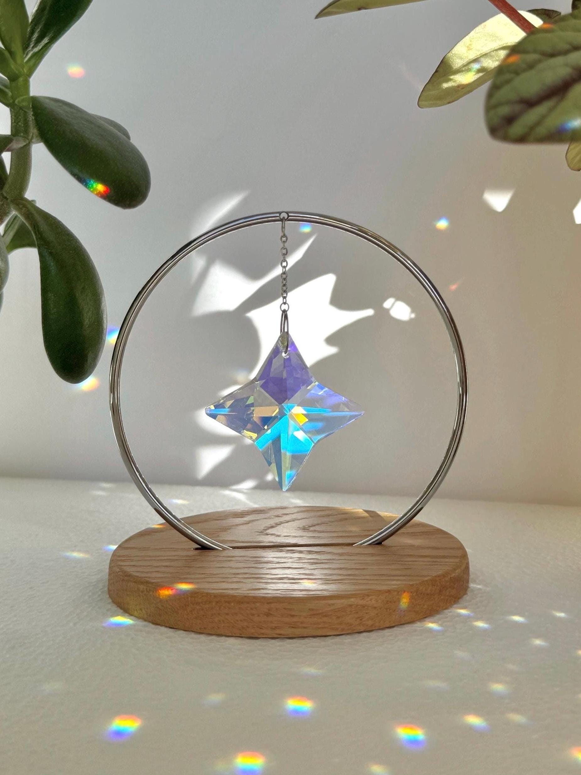 A minimalist Heim decor piece: The Halo Suncatcher featuring a brilliant Kristall-Stern (crystal star) suspended in a silver ring. The star catches the light, scattering vibrant rainbows across a white surface next to a warm wooden base. Wunderschön!