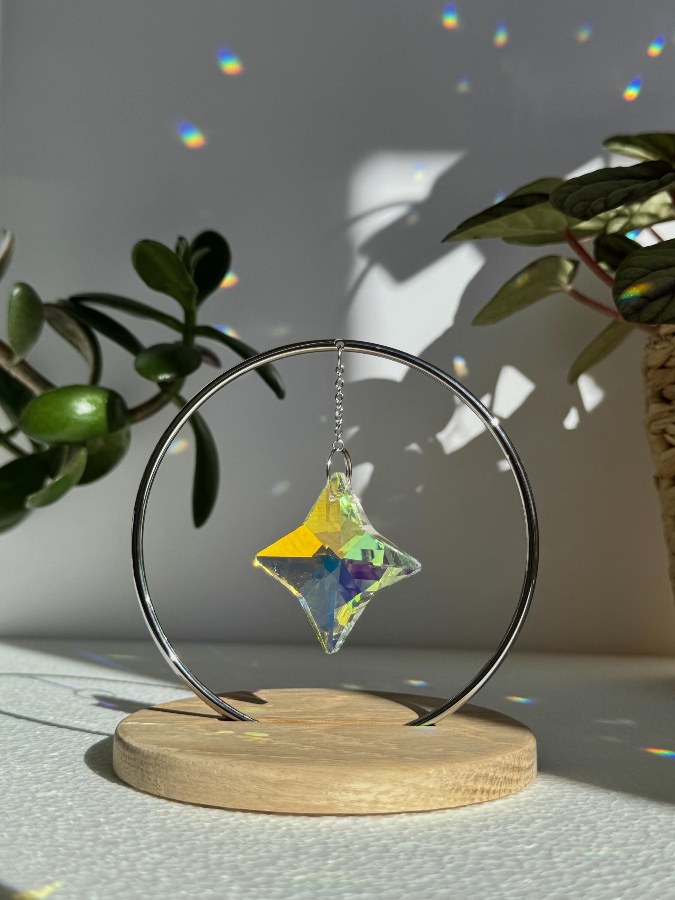 A minimalist Heim decor piece: The Halo Suncatcher featuring a brilliant Kristall-Stern (crystal star) suspended in a silver ring. The star catches the light, scattering vibrant rainbows across a white surface next to a warm wooden base. Wunderschön!