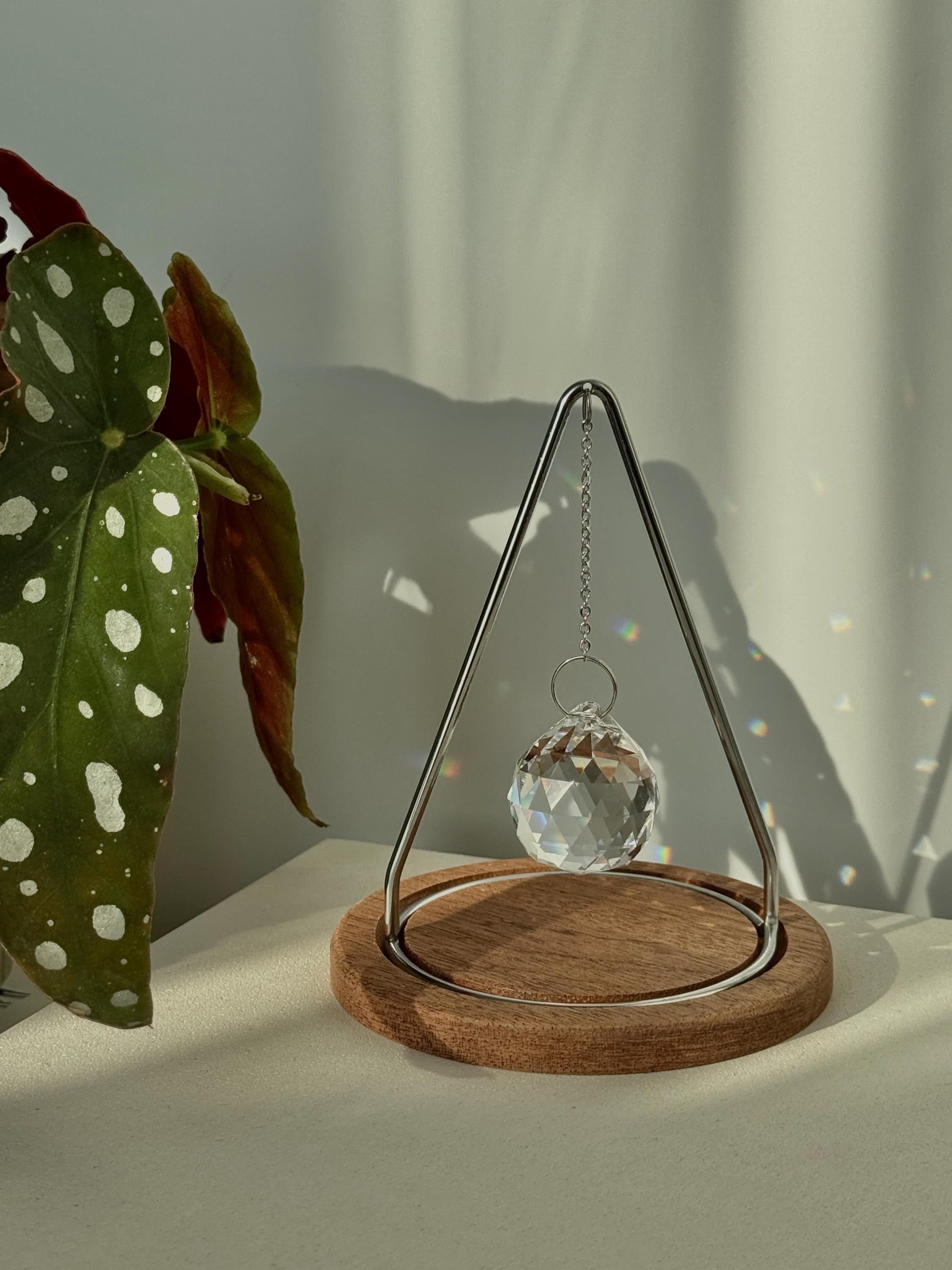 Modern tabletop suncatcher named "The Triangle" featuring a faceted crystal ball prism suspended within a stainless steel frame on a circular redwood base, casting vibrant rainbows on a white surface.