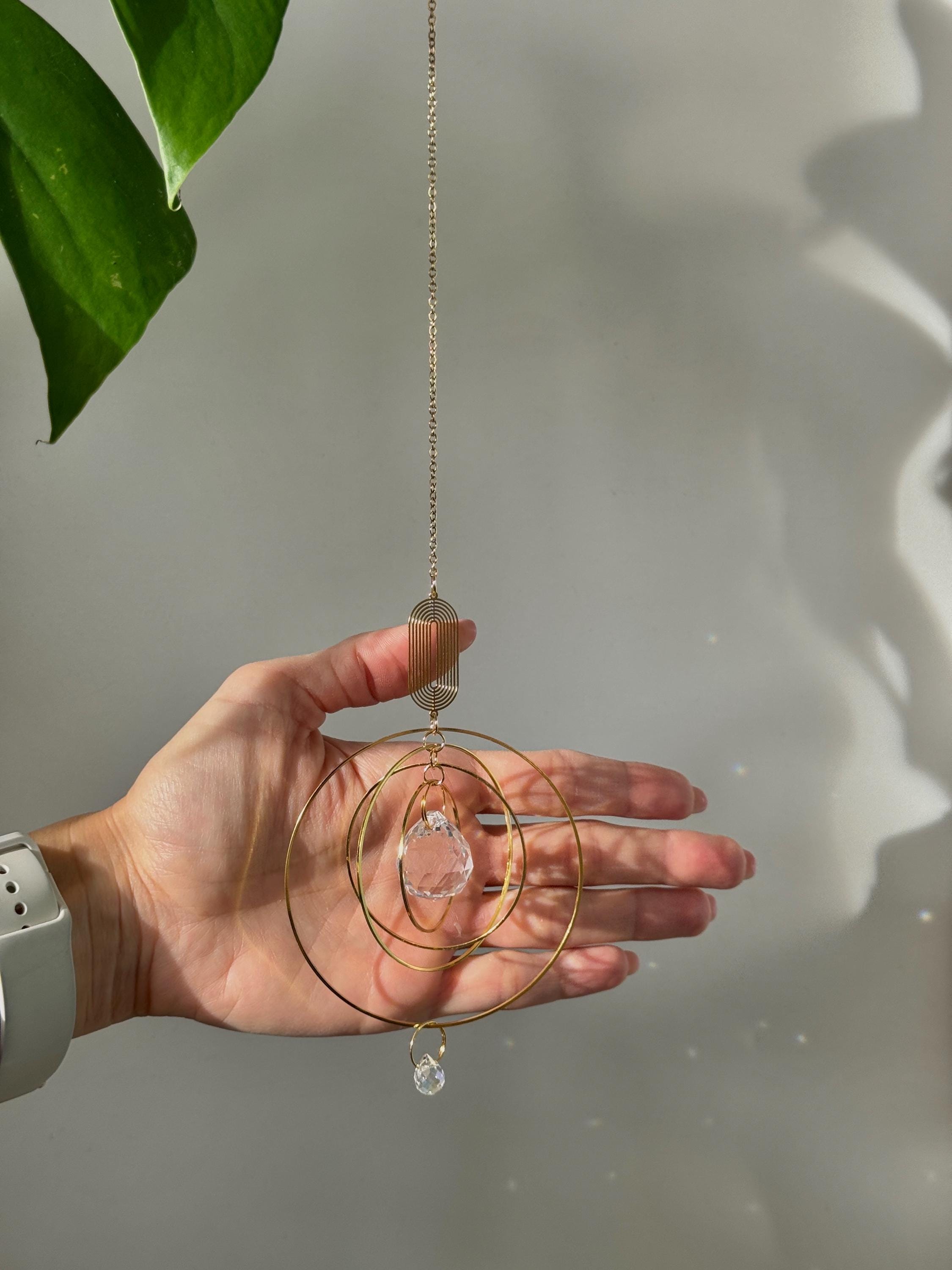 A hand holding a celestial gold window suncatcher (Sonnenfänger) to show the 8cm scale, featuring concentric rings and a crystal prism casting soft light reflections.