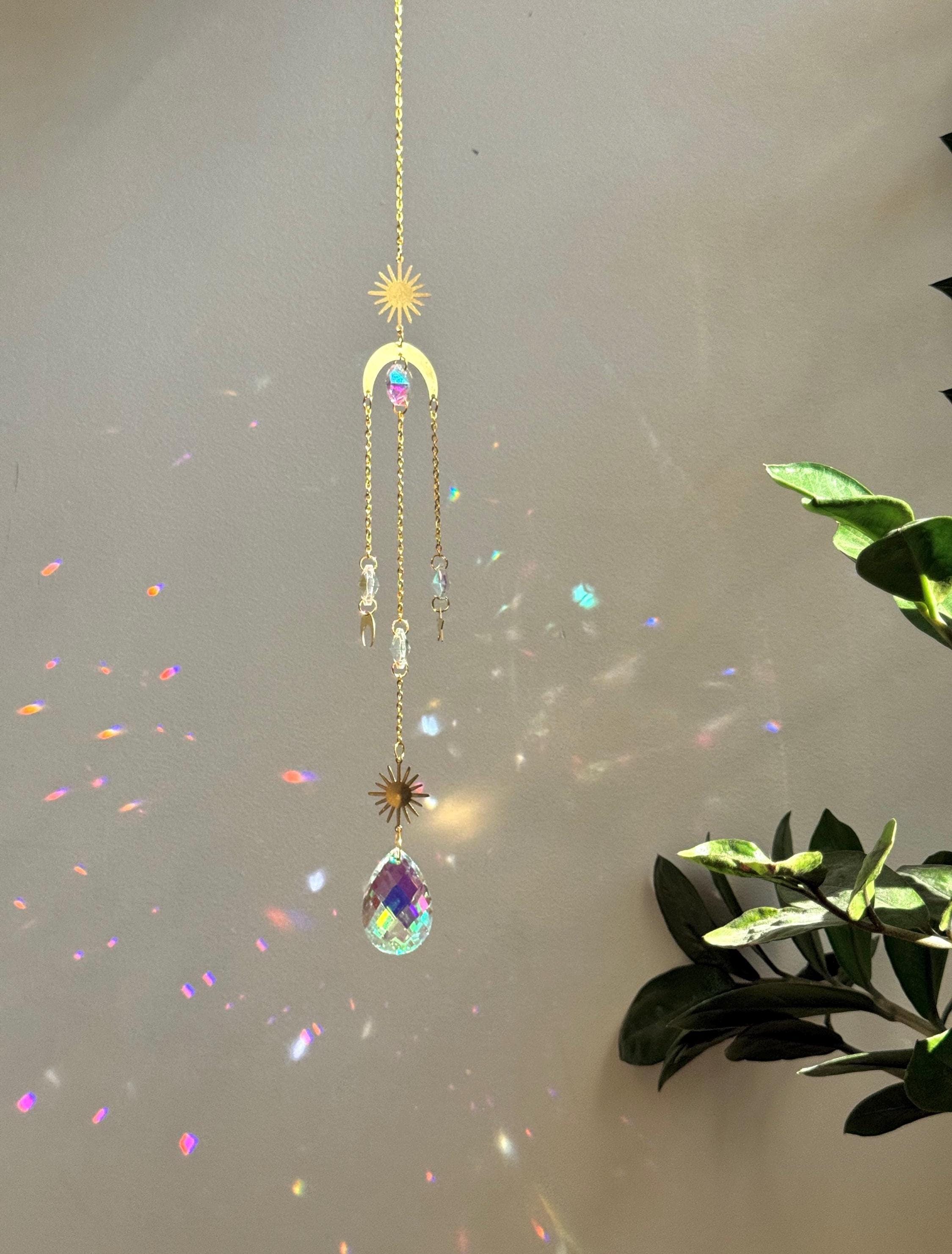 The 'Cosmic Balance' suncatcher, 47cm long, featuring a gold celestial geometric frame and a large 4cm crystal prism, hanging in a bright window and casting wide rainbow patterns.