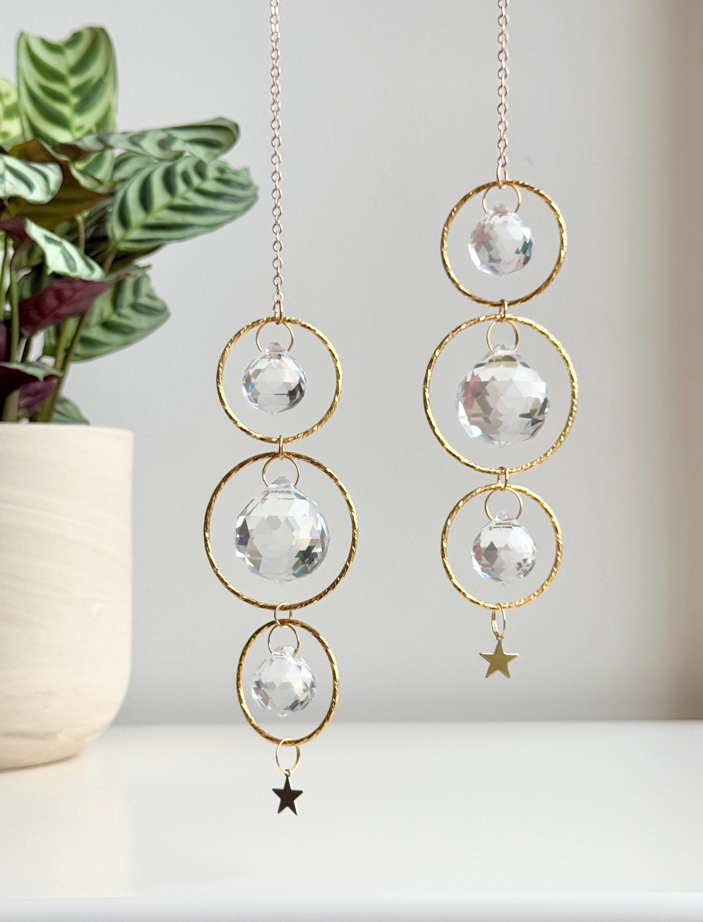 A close-up of the 'Triple Harmony' Suncatcher showing three interlocking gold hoops, each suspending a high-quality faceted glass prism. The central crystal is larger (40mm) than the top and bottom ones (20mm), creating a balanced, tiered effect. The sunlight transforms the room into a canvas of shimmering spectral light and "Sternenstaub" (stardust) reflections. A sharp, rhythmic shadow of the celestial design is cast on the wall behind, highlighting the boho-chic aesthetic.