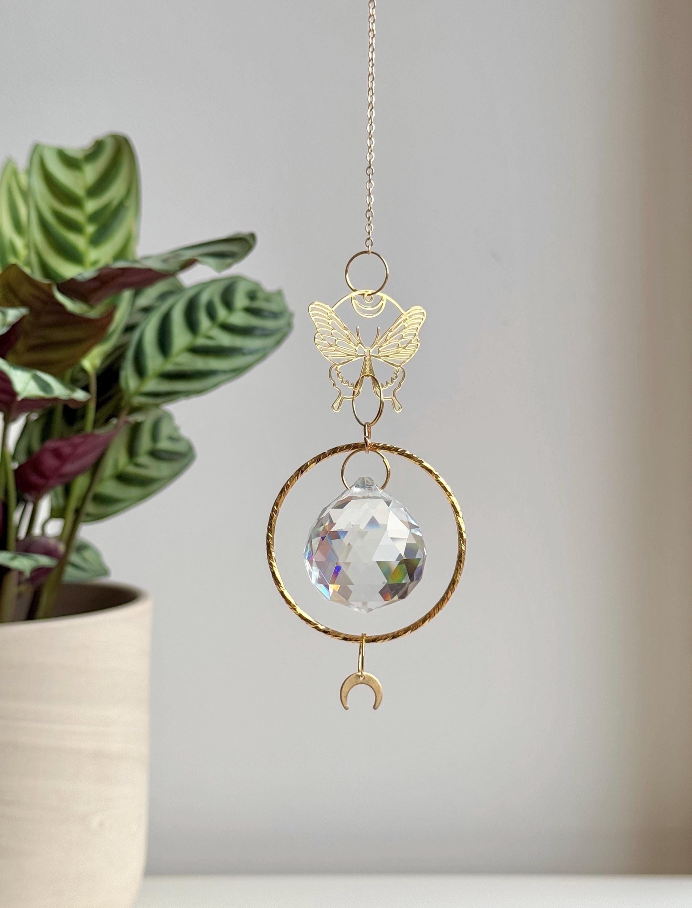 A gold-tone 'Butterfly Effect' Suncatcher features a delicate, etched butterfly charm at the top, followed by a large, circular brass frame housing a premium 40mm faceted crystal prism. A small, downward-facing crescent moon dangles at the very bottom. Direct sunlight illuminates the glass, scattering a magical "Meer von Regenbögen" (sea of rainbows) and "Sternenstaub" (stardust) across the warm, neutral wall. The suncatcher casts a sharp, elegant shadow of the butterfly and moon motifs, creating a serene s