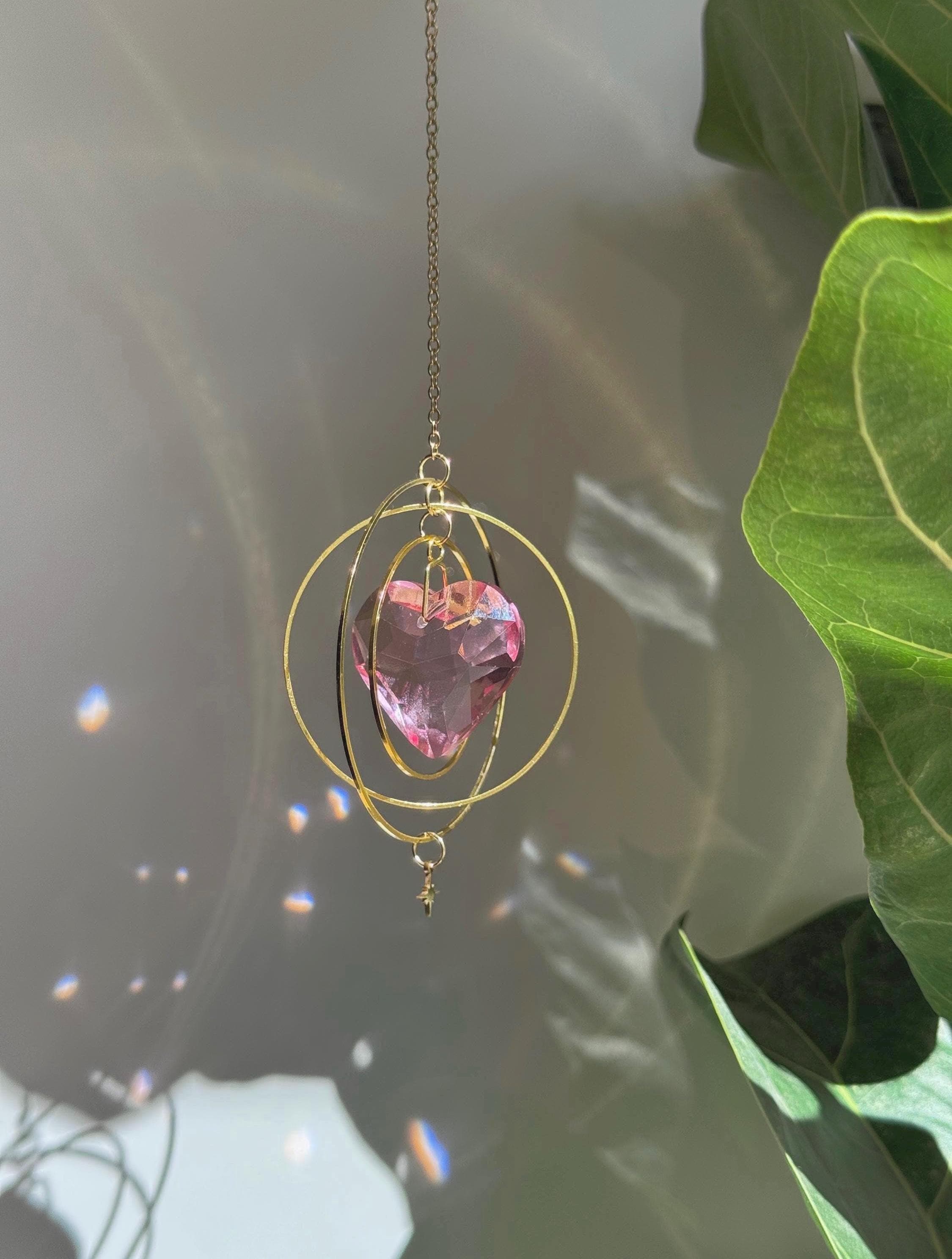 Sunkissed Heartbeat crystal suncatcher featuring a pink-gold faceted heart prism. Handmade celestial window decor with gold rings casting rainbow light flares (Sonnenfänger).
