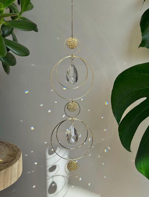 Symmetry in Light – Handmade geometric suncatcher with stainless steel rings and faceted crystal droplets. Celestial window hanging casting rainbows on a wall.