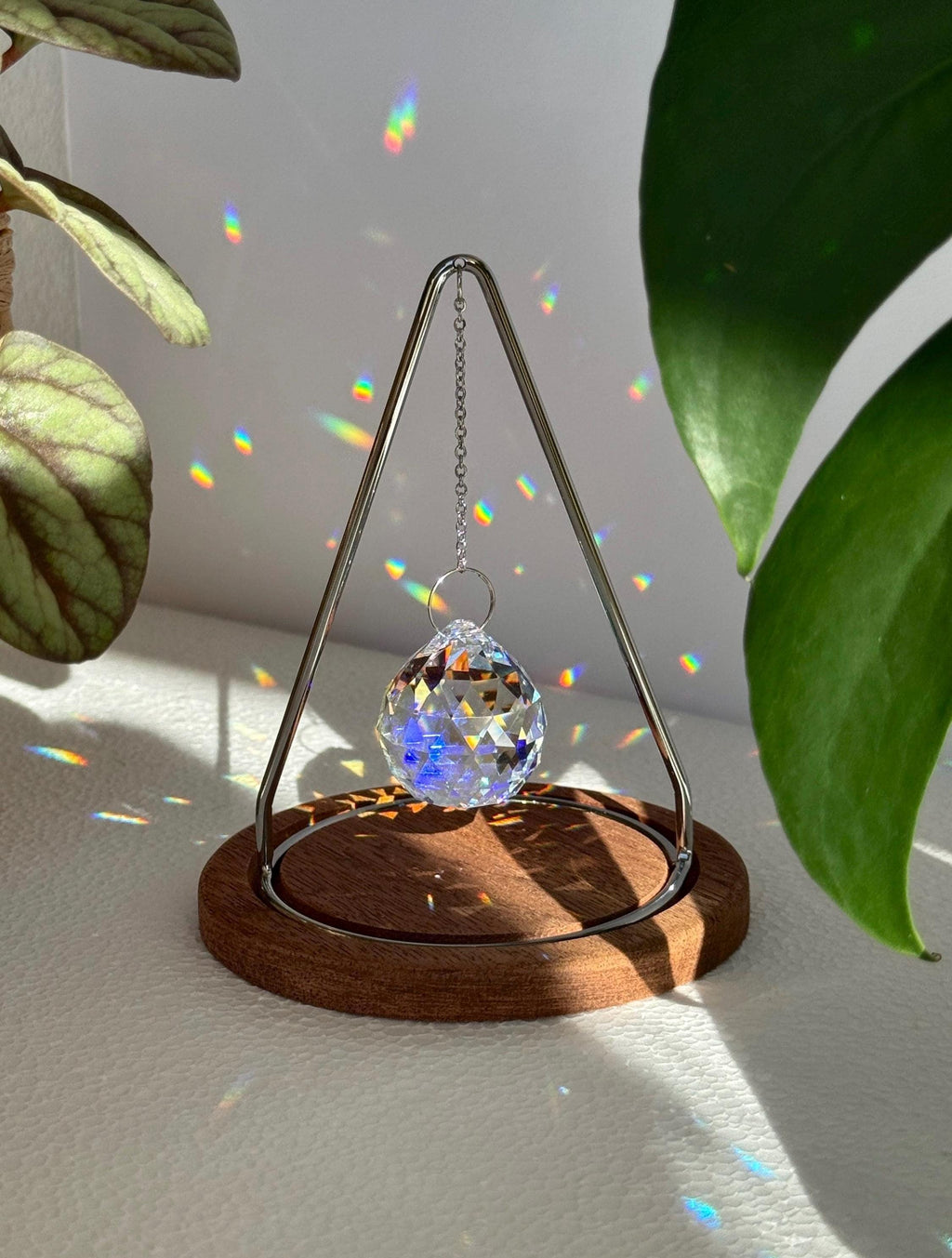 Modern tabletop suncatcher named "The Triangle" featuring a faceted crystal ball prism suspended within a stainless steel frame on a circular redwood base, casting vibrant rainbows on a white surface.