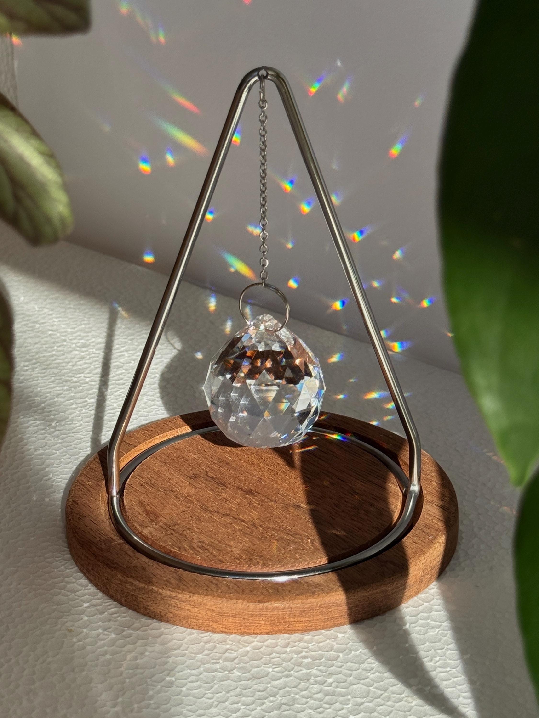 Modern tabletop suncatcher named "The Triangle" featuring a faceted crystal ball prism suspended within a stainless steel frame on a circular redwood base, casting vibrant rainbows on a white surface.