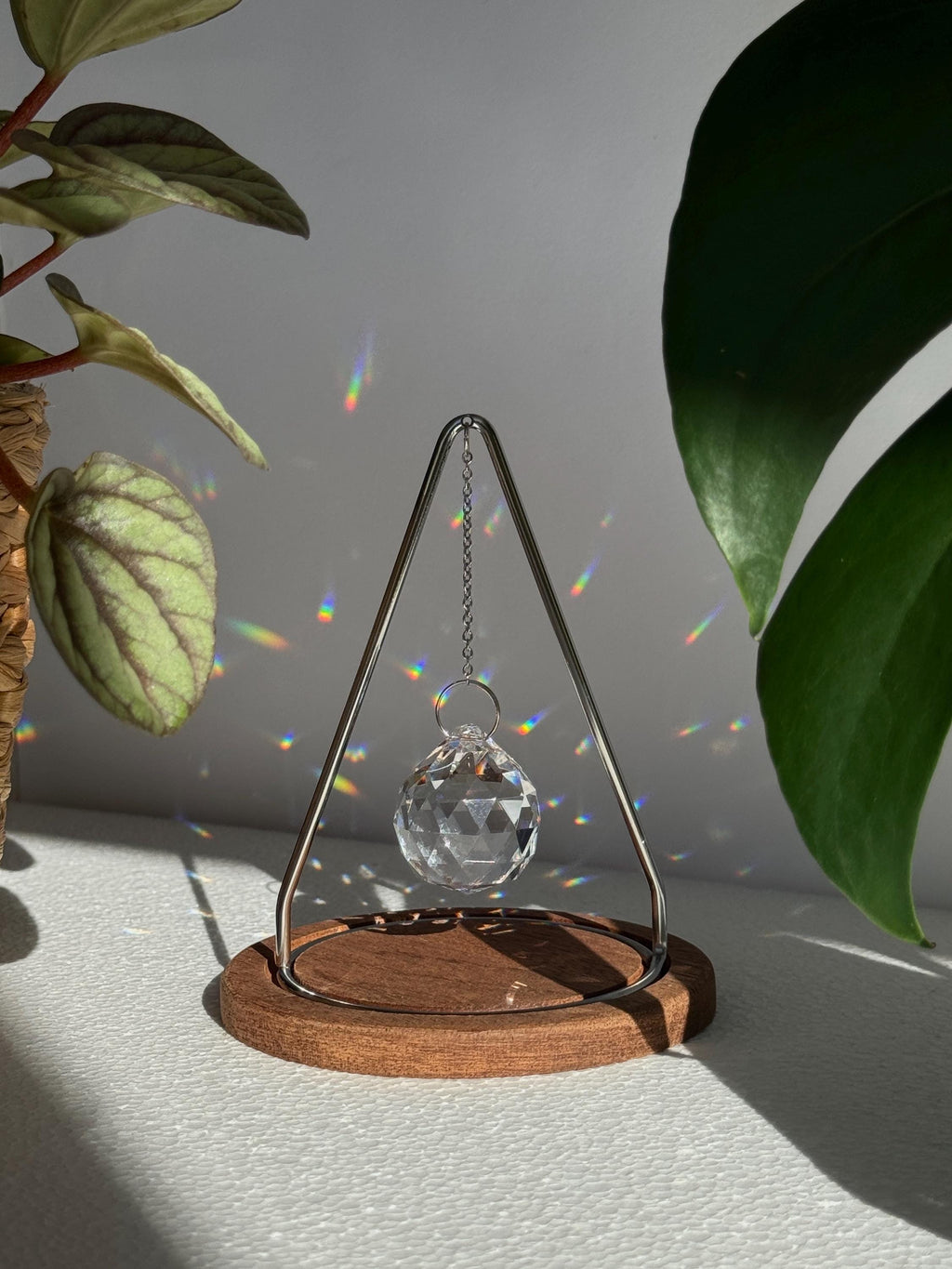 Modern tabletop suncatcher named "The Triangle" featuring a faceted crystal ball prism suspended within a stainless steel frame on a circular redwood base, casting vibrant rainbows on a white surface.