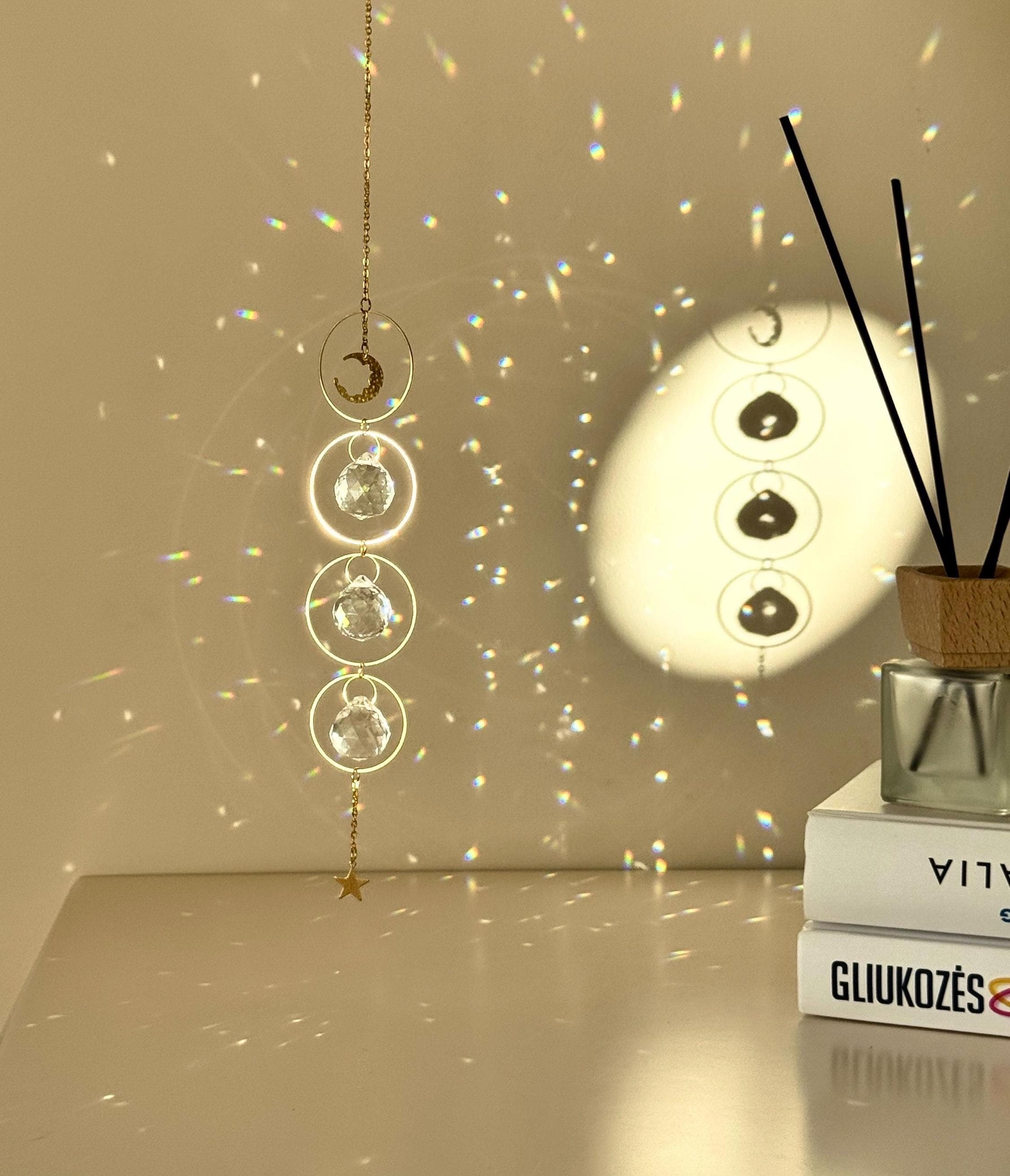 A gold-tone 'Serenity' celestial suncatcher hangs against a warm, neutral wall, featuring three circular brass hoops. The top hoop holds a delicate crescent moon, while the bottom three each frame a 20mm faceted crystal prism that ends in a small gold star. Bright sunlight creates a magical "Meer von Regenbögen" (sea of rainbows) and shimmering light speckles across the surface. To the right, a reed diffuser and stacked books create a sense of Gemütlichkeit (coziness), while the suncatcher casts a soft, rhy