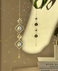 A vertical gold-tone geometric suncatcher hangs elegantly against a cream-colored wall, casting a dazzling "Meer von Regenbögen" (sea of rainbows) across the room. The piece features alternating small and large diamond-shaped brass frames, with two faceted 2cm crystal prisms suspended in the center. Sunlight passes through the glass, creating vibrant spectral flares that dance like "Sternenstaub" (stardust). To the right, a lush green Monstera leaf and a stack of books add a modern, boho-chic vibe. The over