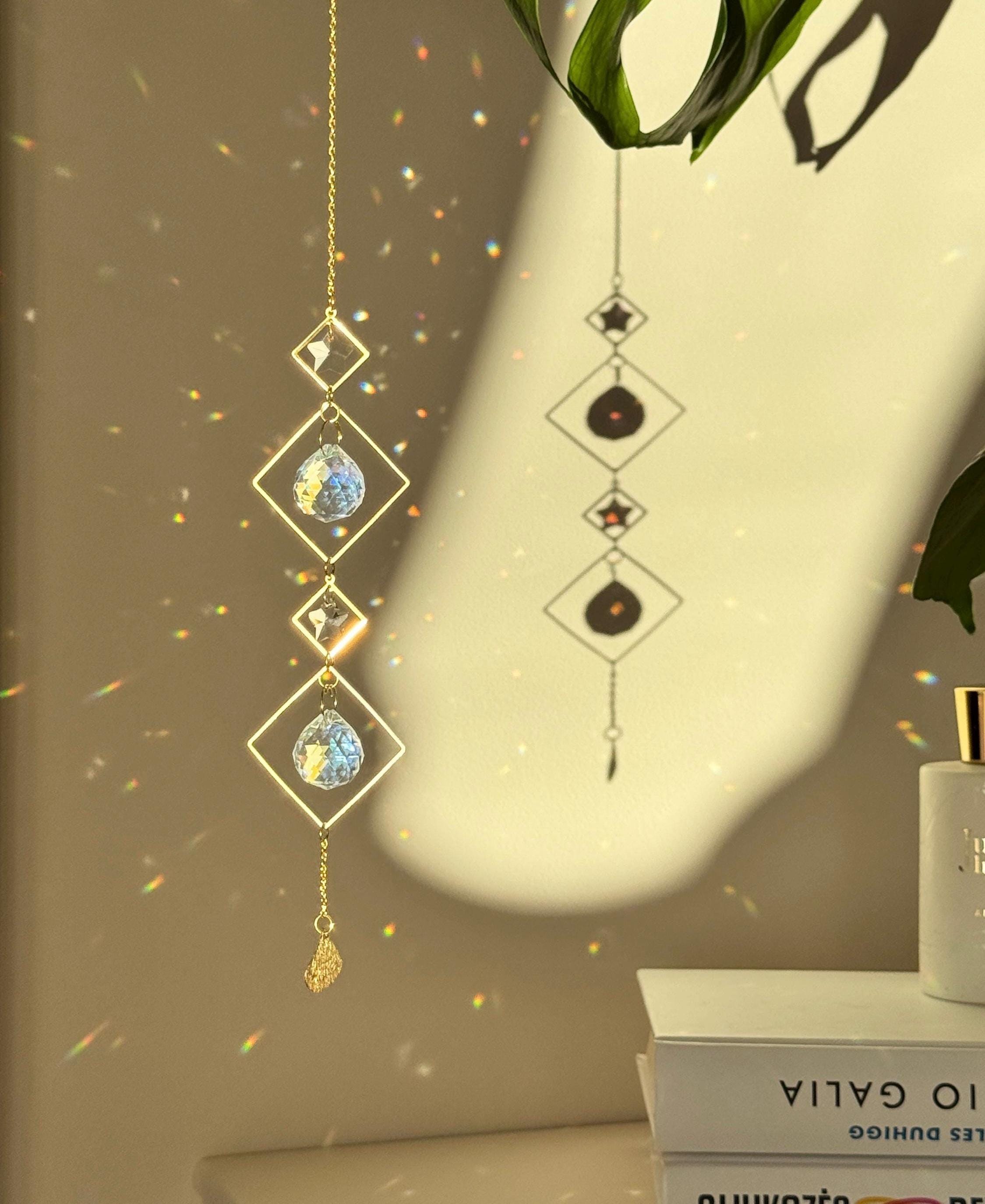 A vertical gold-tone geometric suncatcher hangs elegantly against a cream-colored wall, casting a dazzling "Meer von Regenbögen" (sea of rainbows) across the room. The piece features alternating small and large diamond-shaped brass frames, with two faceted 2cm crystal prisms suspended in the center. Sunlight passes through the glass, creating vibrant spectral flares that dance like "Sternenstaub" (stardust). To the right, a lush green Monstera leaf and a stack of books add a modern, boho-chic vibe. The over