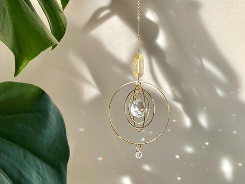 Celestial boho window suncatcher (Sonnenfänger Fenster) with gold concentric rings and a crystal prism, scattering magical rainbows (Regenbogen) across a sunlit wall next to a Monstera leaf.