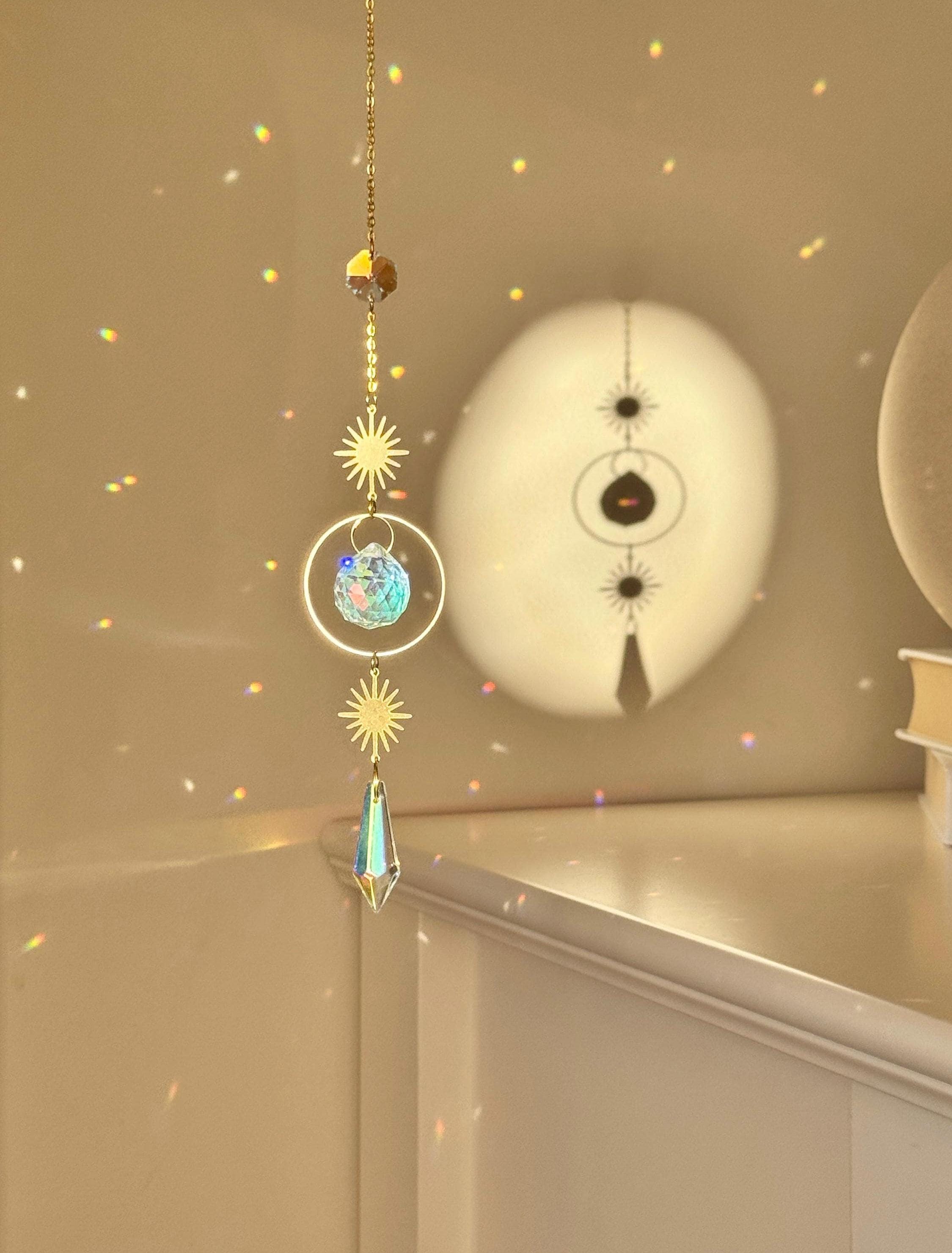 Celestial 'Sunny Days' suncatcher with gold sun charms and two crystal prisms hanging against a warm-toned wall, casting small rainbow light spots across the room.