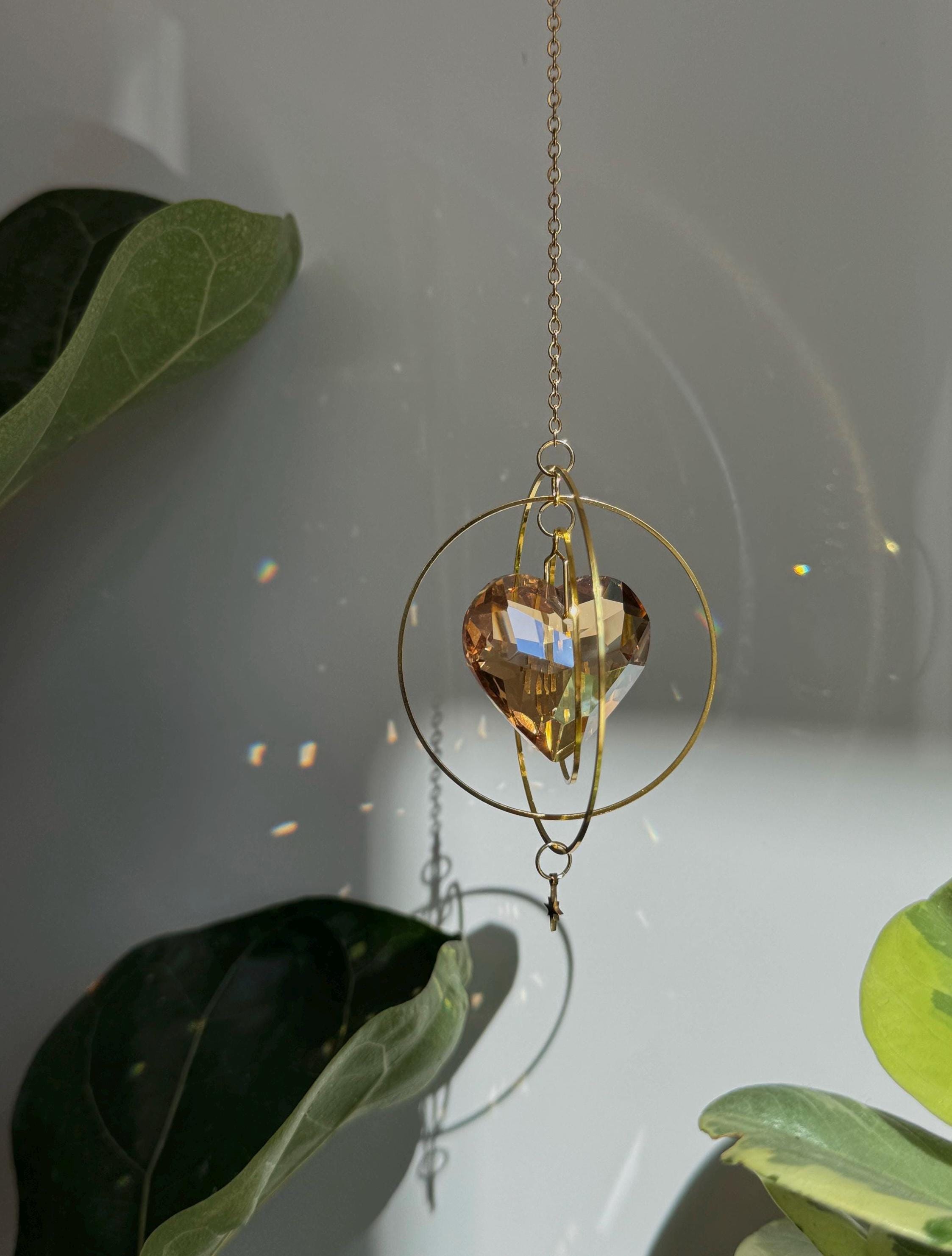Sunkissed Heartbeat crystal suncatcher featuring a warm amber-gold faceted heart prism. Handmade celestial window decor with gold rings casting rainbow light flares (Sonnenfänger).