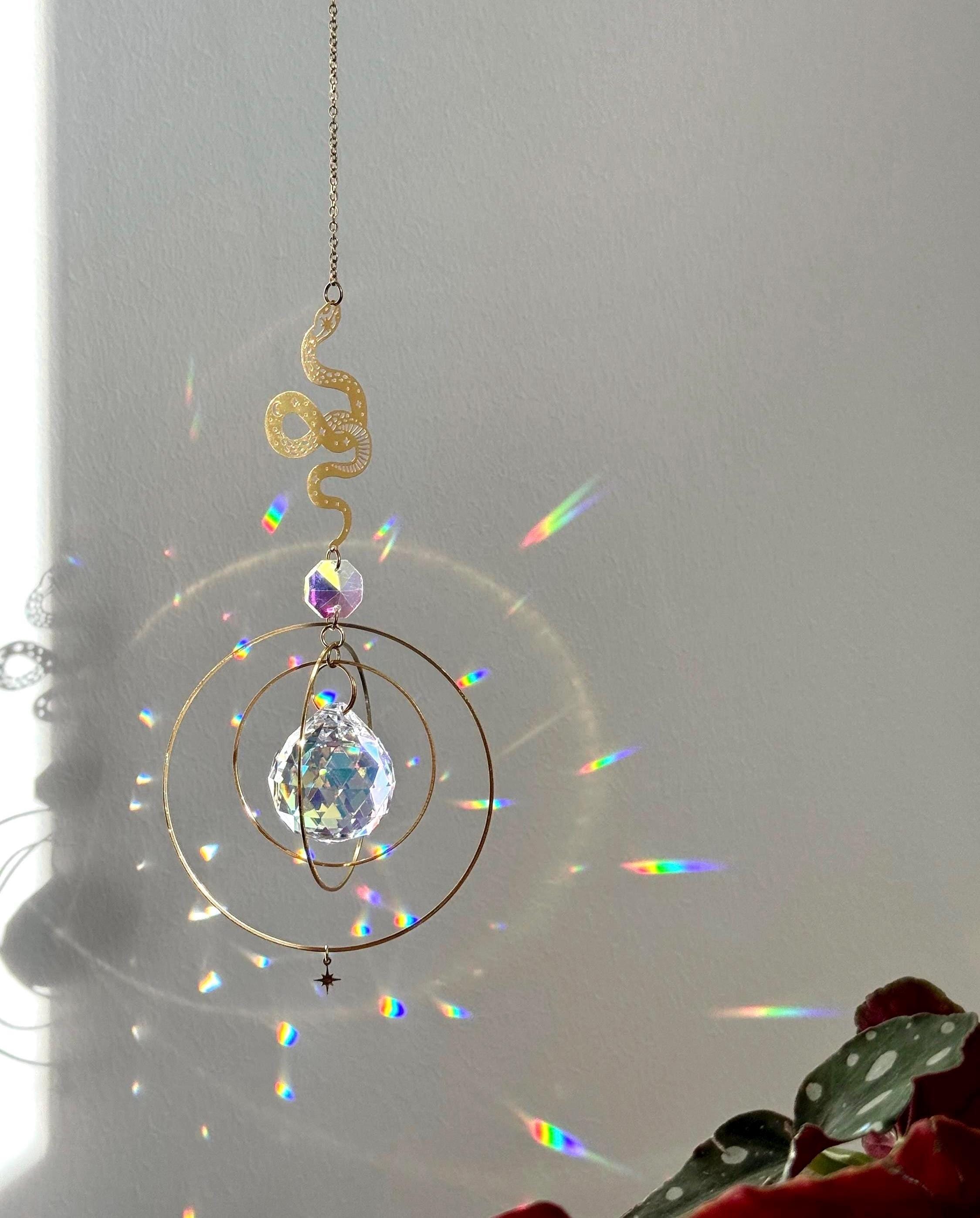 Handcrafted Sunkissed Heartbeat suncatcher with a faceted crystal heart and gold orbital rings casting vibrant rainbows on a white wall near a green plant.