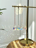 Four crystal prism car suncatchers in different sizes hanging from a gold jewelry stand, casting rainbow light reflections on a white wall next to a green plant.