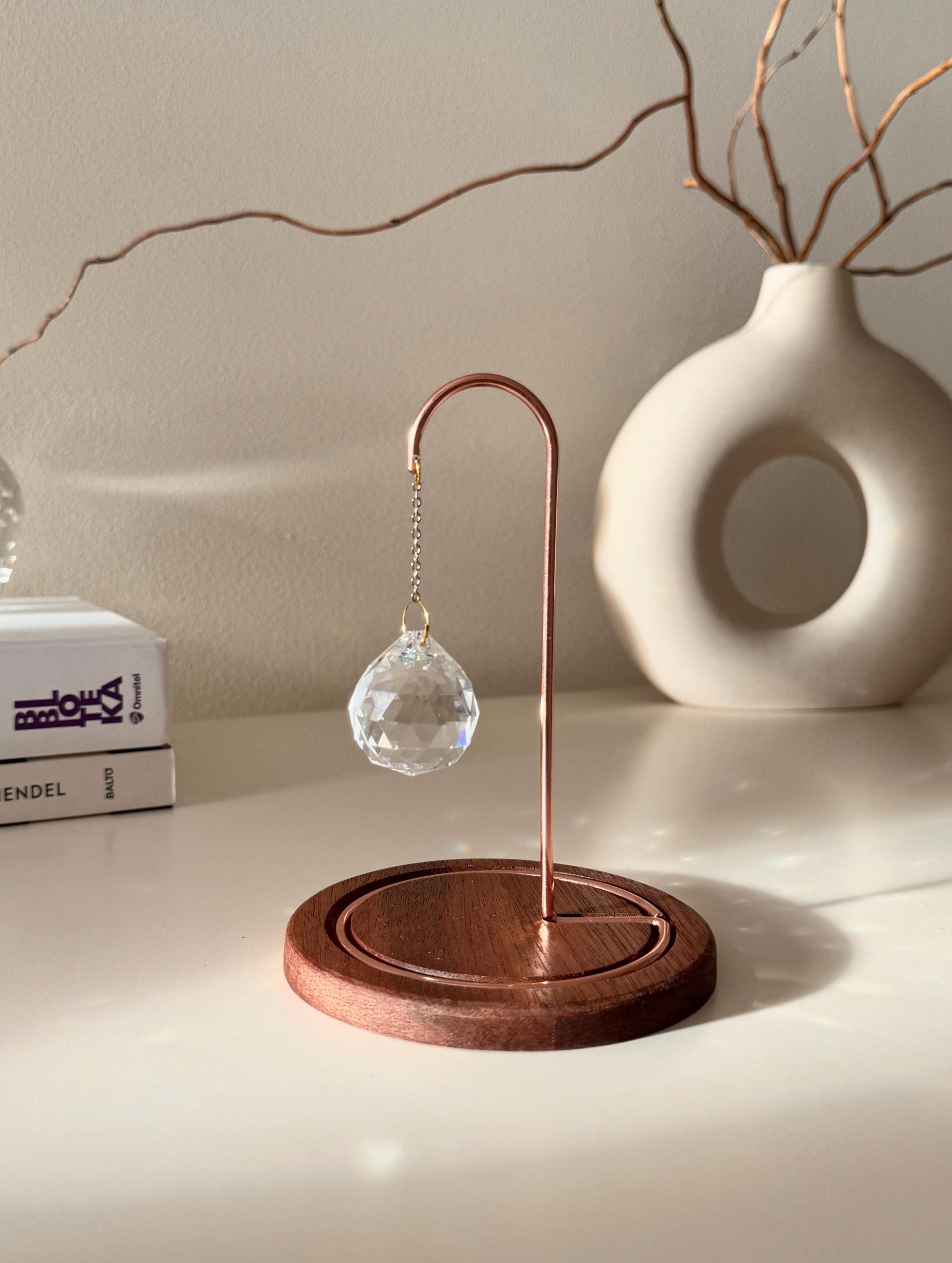 A close-up photograph of a clear, multi-faceted crystal suncatcher prism hanging from an ornate rose gold metal hook on a dark wooden base. The suncatcher casts multiple rainbow-colored refractions of light across the white tabletop, a stack of white books on the left, and a textured off-white ceramic vase in the background. Dried branches are visible emerging from the vase against a light gray wall.
