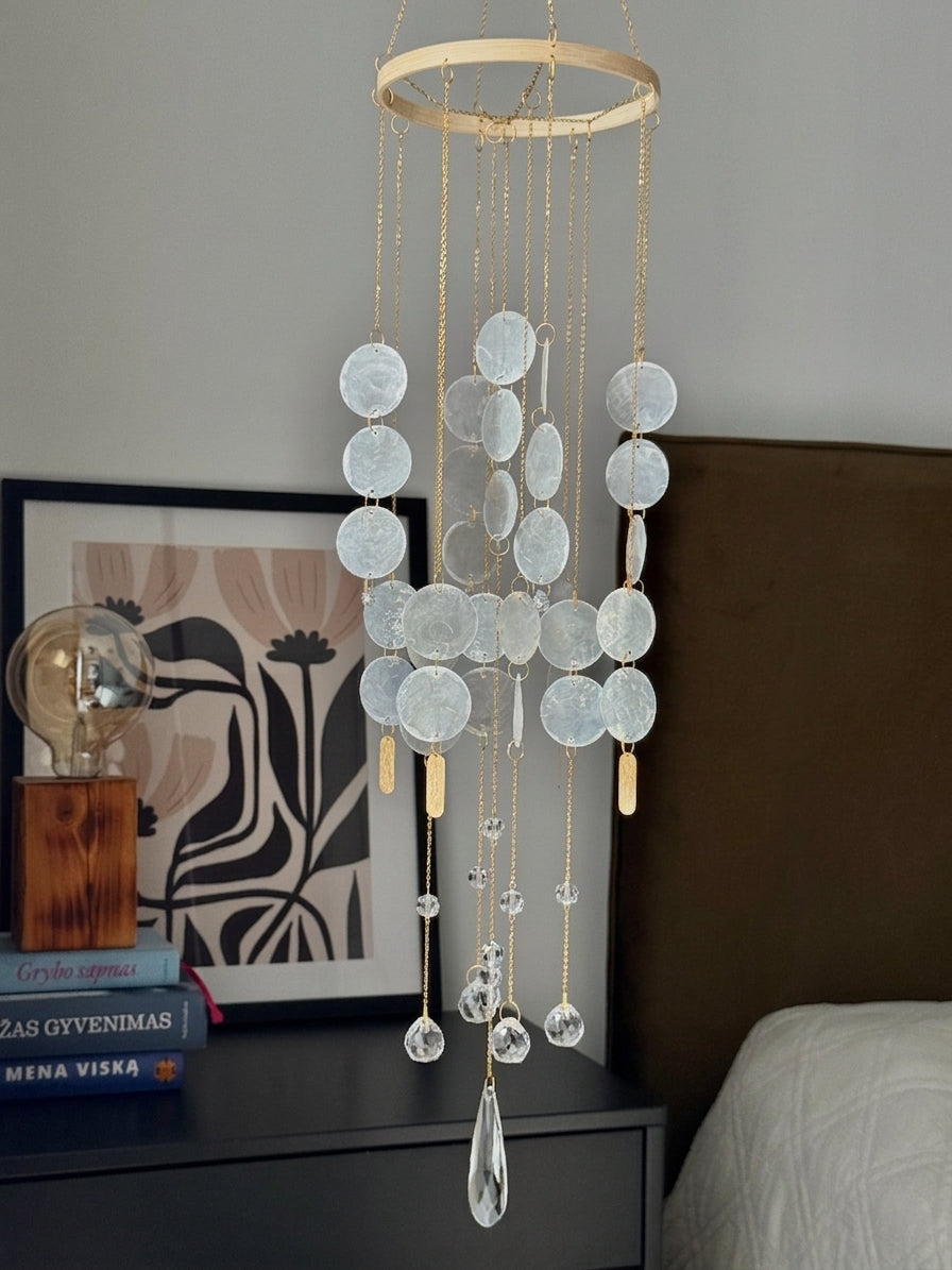 A hanging wind chime with circular clear glass discs on a gold-toned ring frame, suspended in a bedroom setting above a dark bedside table.